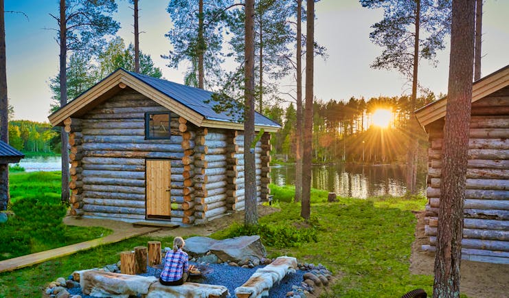 Arctic Retreat cabin exteriors and communal firepit overlooking river, sun setting amongst tall trees