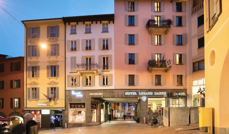 Hotel Lugano Dante exterior, pink hotel building, shop fronts, sunset, town square
