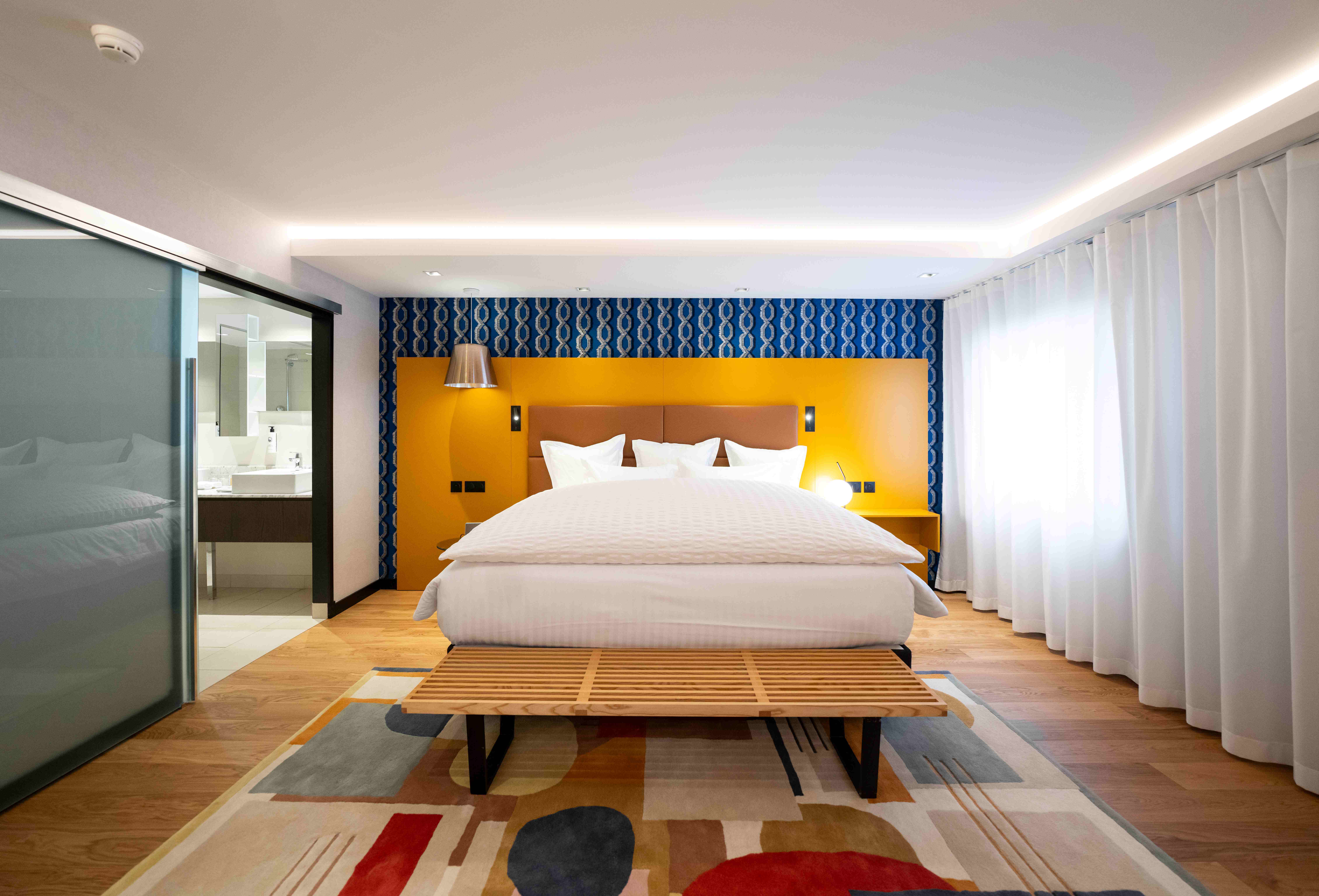 Assiette Champenoise junior suite bedroom with yellow and blue colour scheme, large bed, patterned rug, and white curtains covering the large window