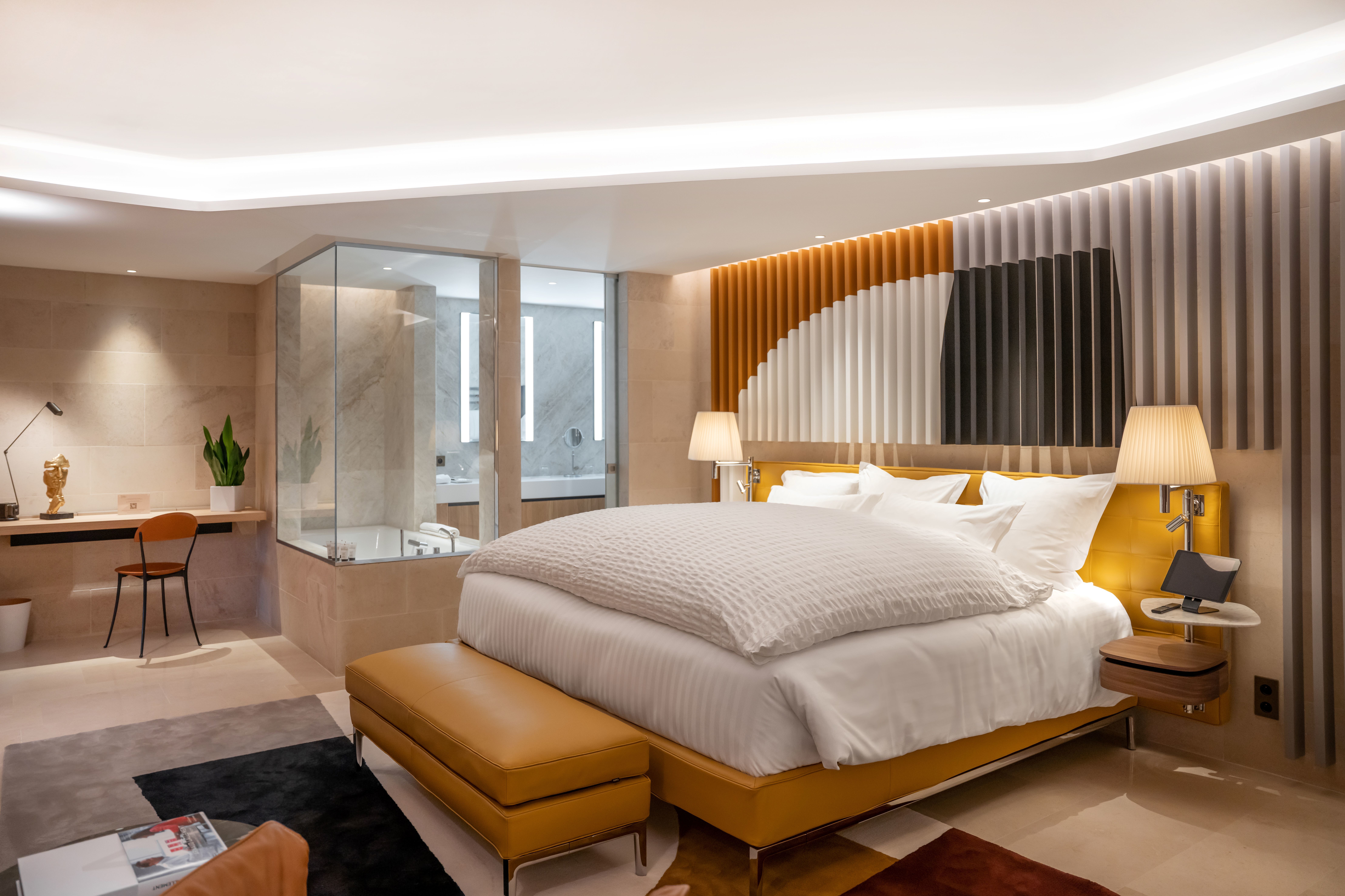 Assiette Champenoise suite room with ochre and grey colour scheme, large bed, desk area, glass-walled bathroom, and lighting around the tops of the walls