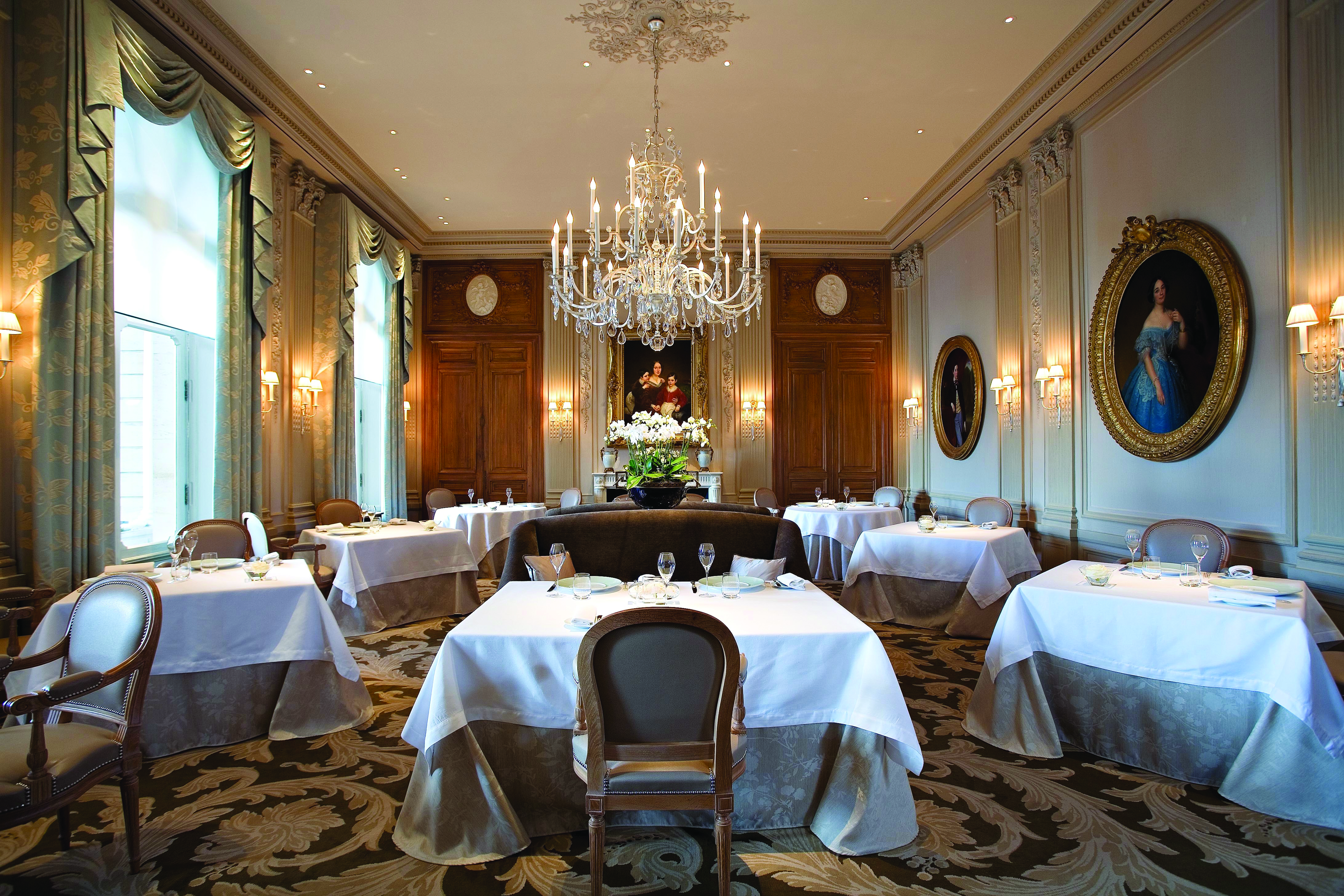 Dining room with large crystal chandelier, tables and chairs set up and gold framed art work on the walls