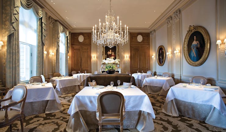 Dining room with large crystal chandelier, tables and chairs set up and gold framed art work on the walls
