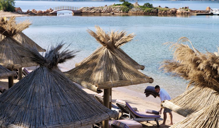 Grand Hotel de Cala Rossa Corsica beach with sun loungers and wooden umbrellas and bridge to a small island