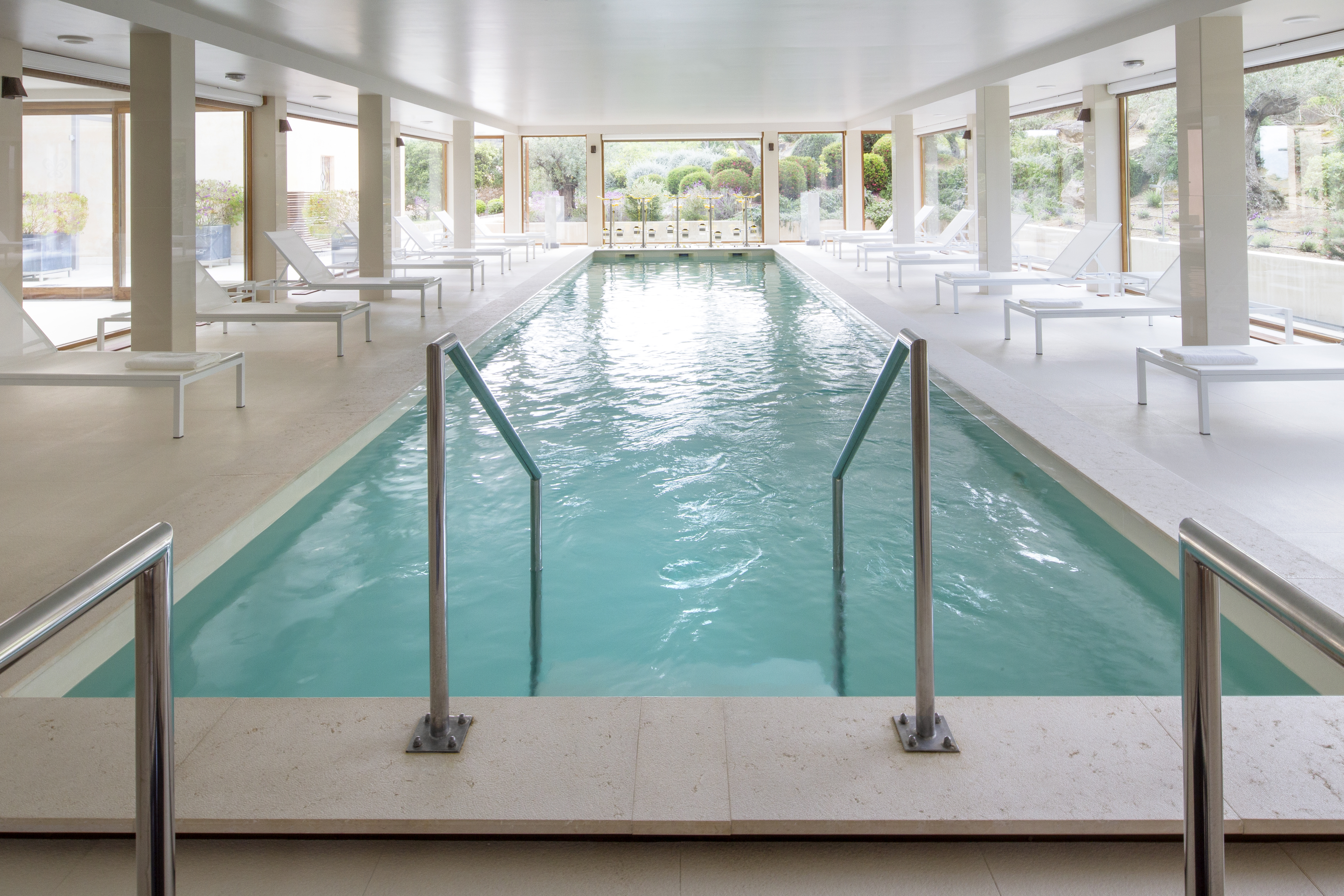 Indoor swimming pool with window pannelled walls and white sun loungers around the edge of the pool