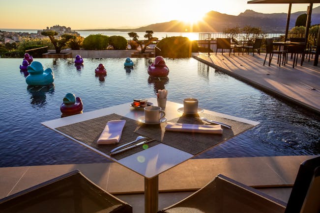 Outdoor pool with dining table set out around the edge, sun setting behind mountains in the background and ducks in the pool