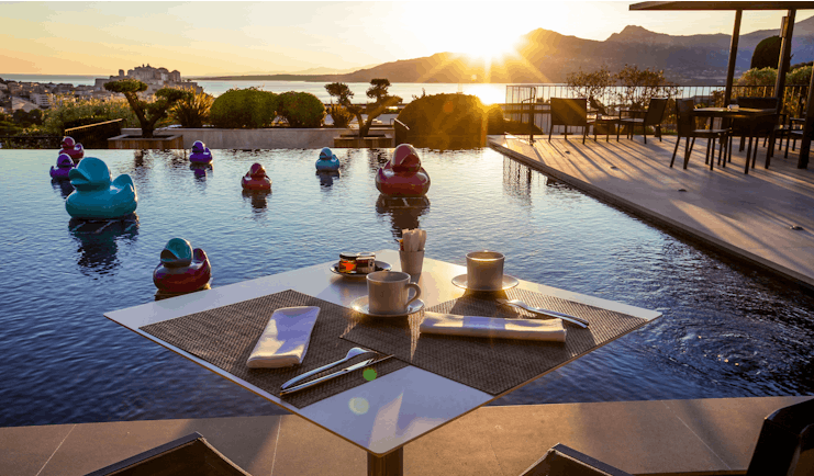 Outdoor pool with dining table set out around the edge, sun setting behind mountains in the background and ducks in the pool