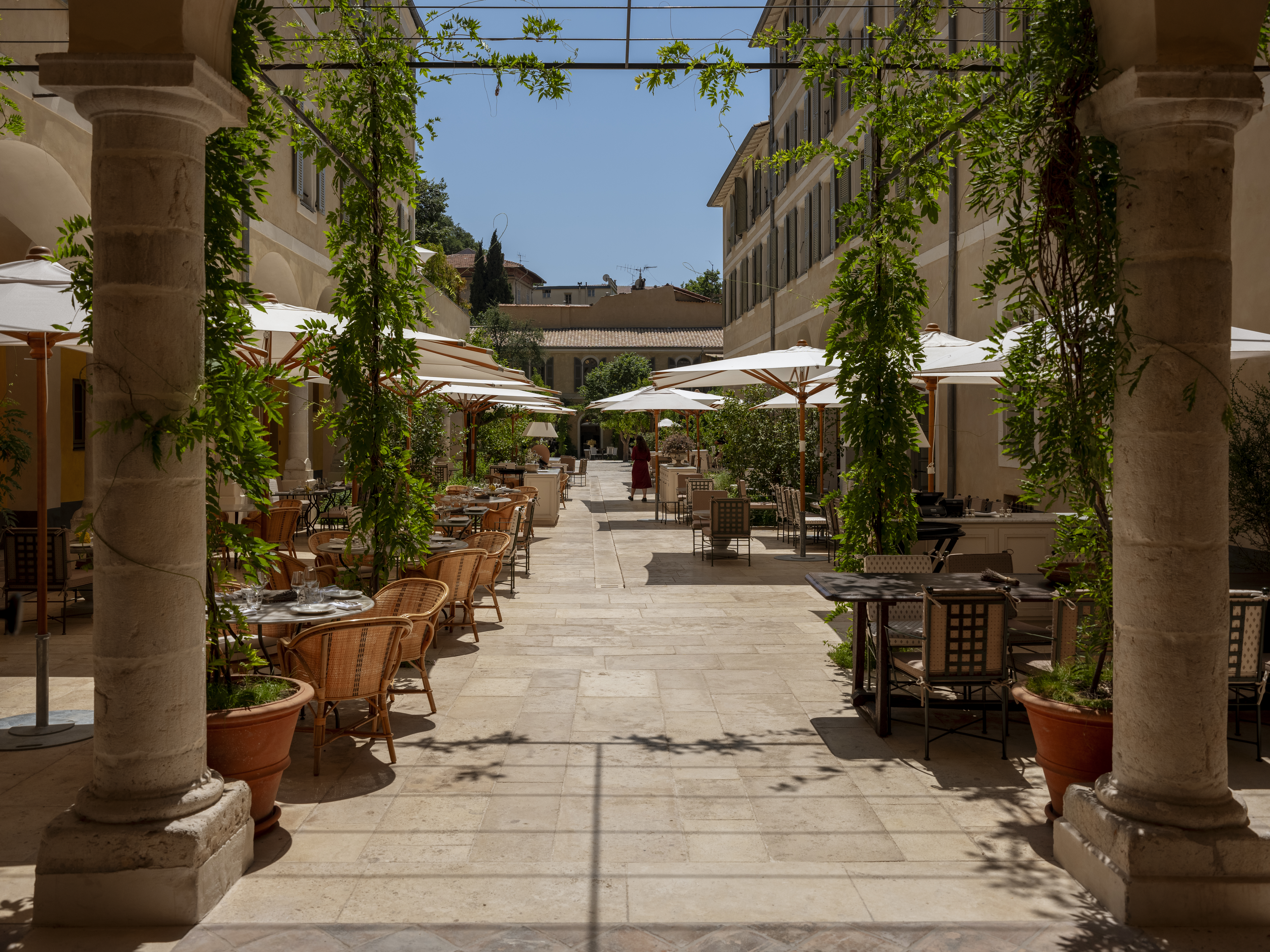 Hotel du Couvent Cloister Courtyard, with ochre hotel buildings on both sides, lined with tables, chairs, and parasols, with winding vines and stone pillars in the foreground