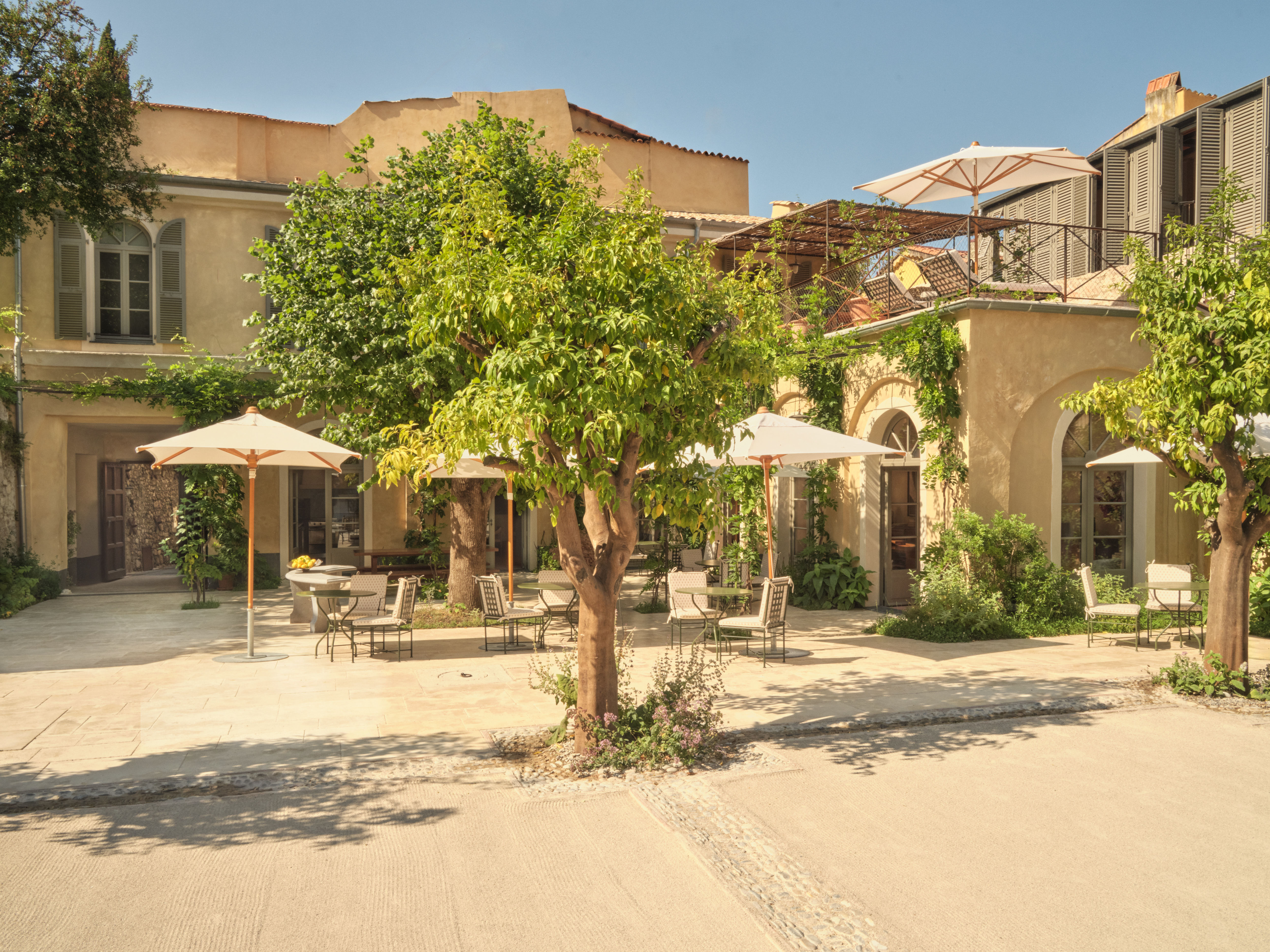 Hotel du Couvent outdoor dining with chairs and tables under parasols and trees, with the arches and shutters of the hotel building in the background