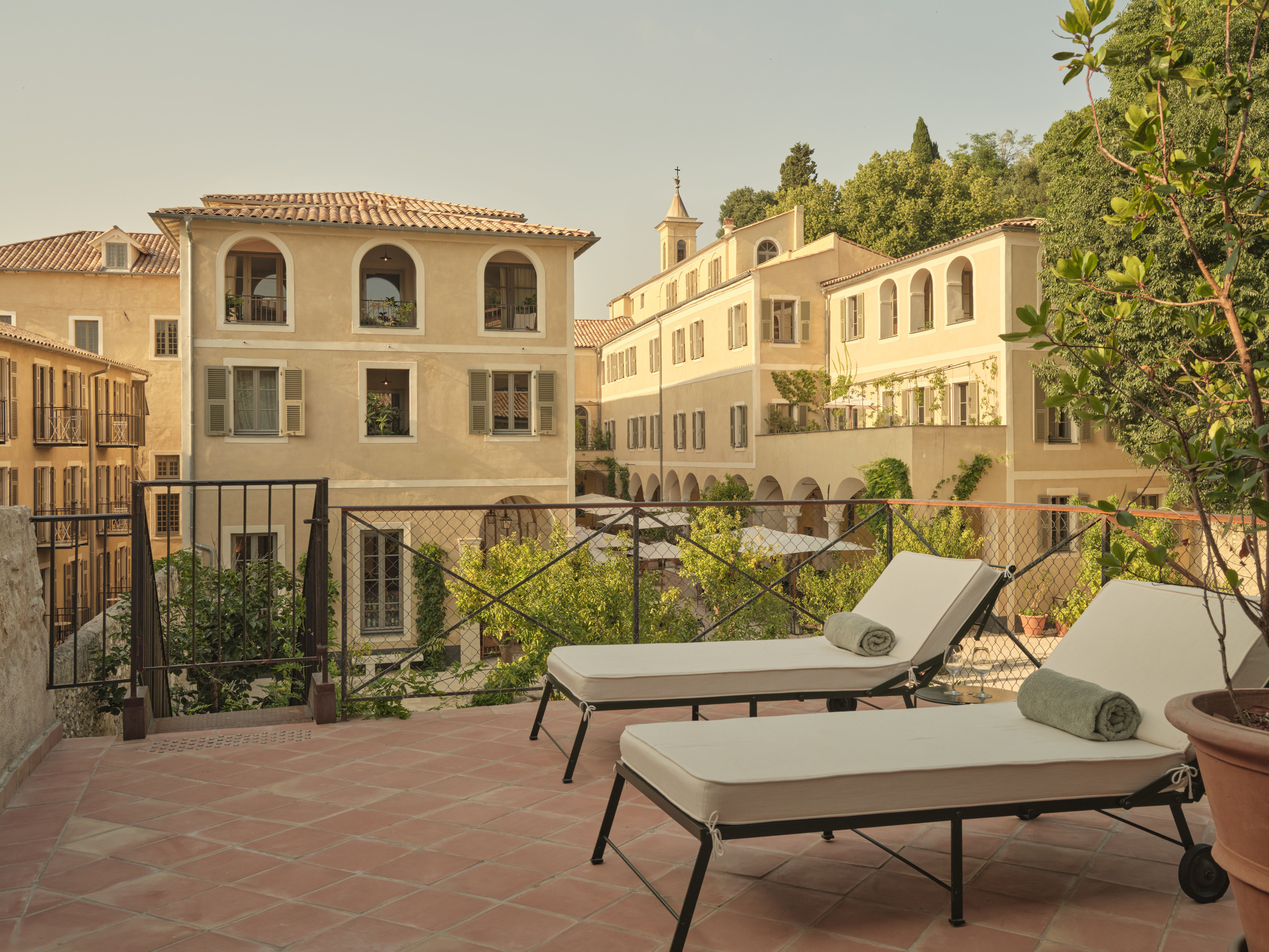 Hotel du Couvent terrace with terracotta floor tiles, warm white sun loungers, and the ochre hotel buildings, with arched windows and tiled roofs and trees in the background