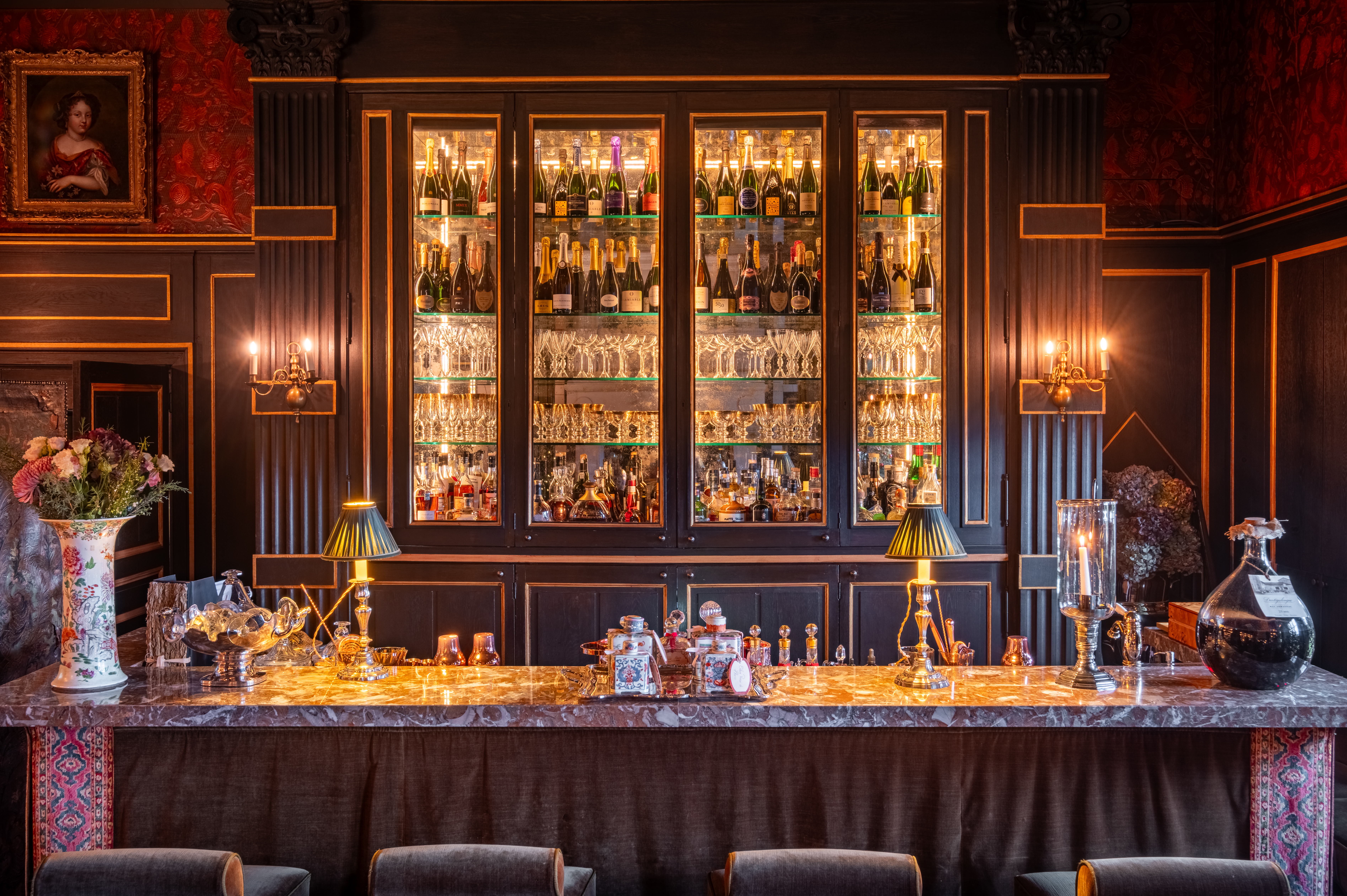 Chateau Louise de la Valliere bar, with lit glass cabinet of bottles, antique portraiture on the walls, flowers in vases, and personal lamps