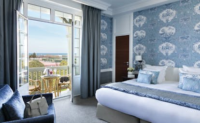 Double room with blue colour scheme, large double bed, and double windows opening onto a small balcony with table and chairs on 