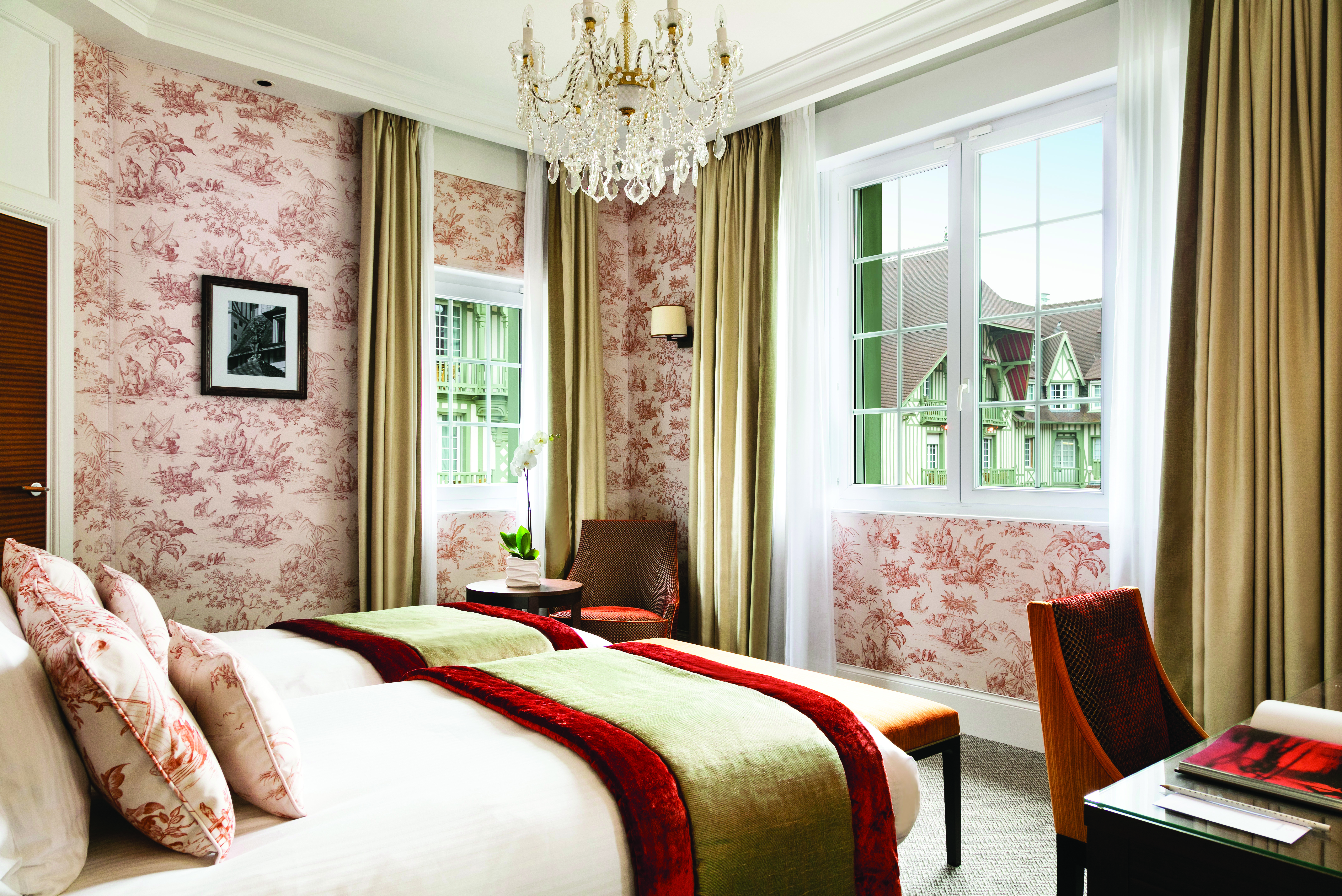 Twin room with two single beds, pink, red and beige colour scheme and two large windows with a glass chandelier hanging from the ceiling