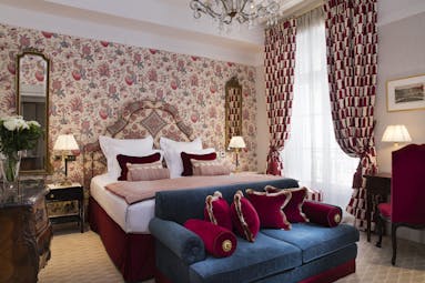 Deluxe room with floral patterned wallpaper, double bed, sofa and draping curtains 