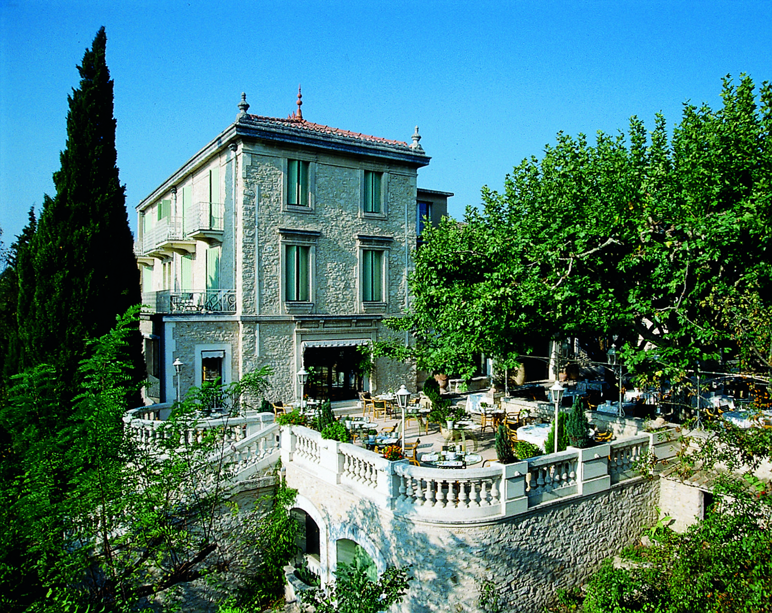 Exterior of the Auberge de Noves in Provence with stone building, large trees surrounding it and a outdoor patio area