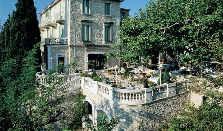 Exterior of the Auberge de Noves in Provence with stone building, large trees surrounding it and a outdoor patio area