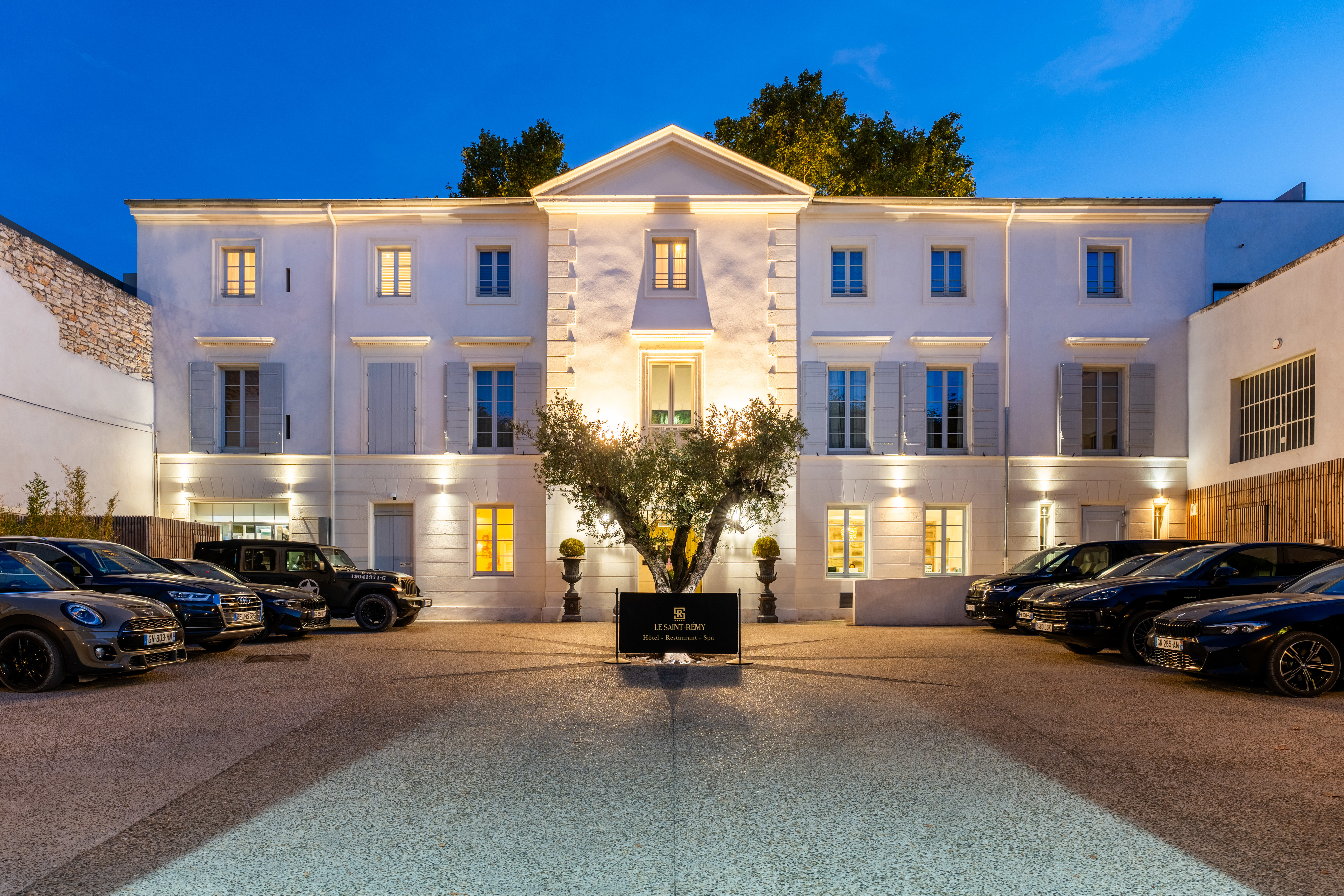 Hotel Le Saint-Remy grand entrance with white stone walls, a symmetrical facade, and a central tree in the parking courtyard