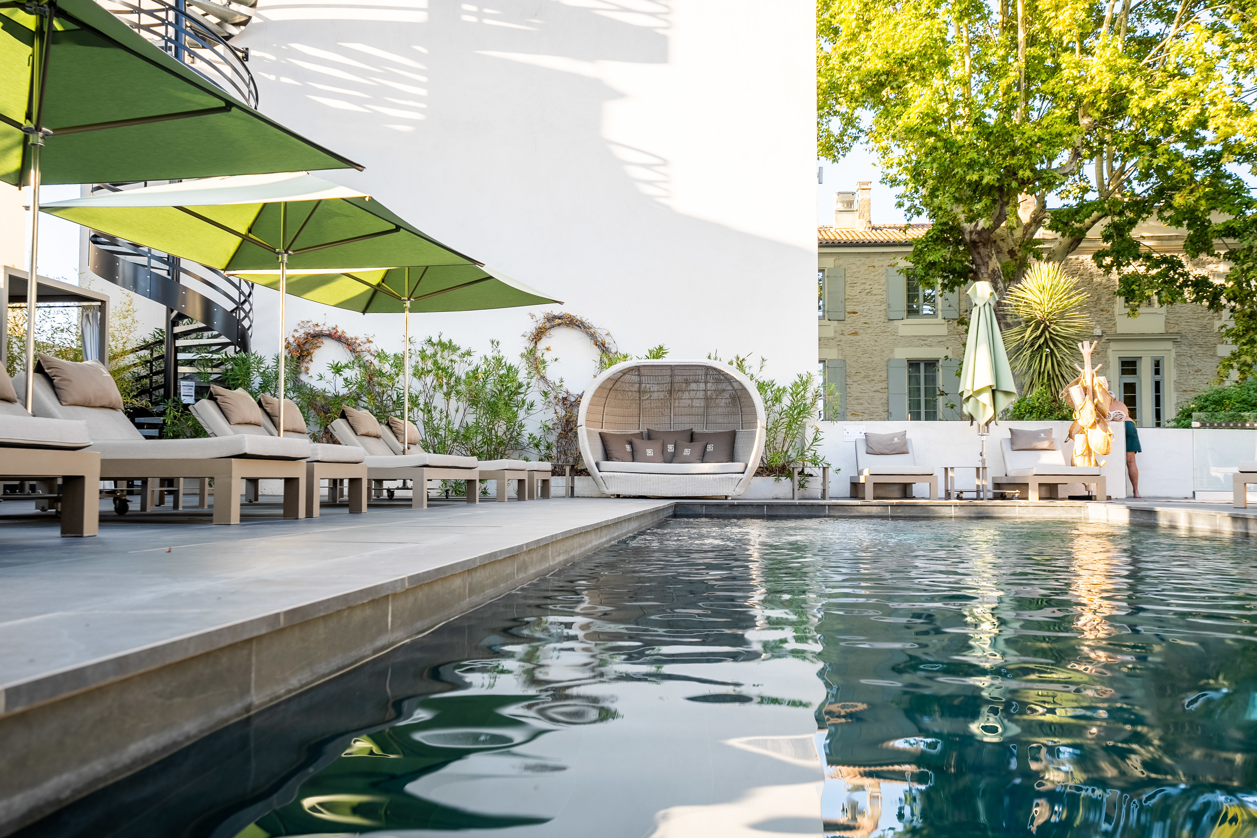 Hotel Le Saint-Remy swimming pool, seen from water-level, with sun loungers under green parasols, daybeds, and tall shady trees