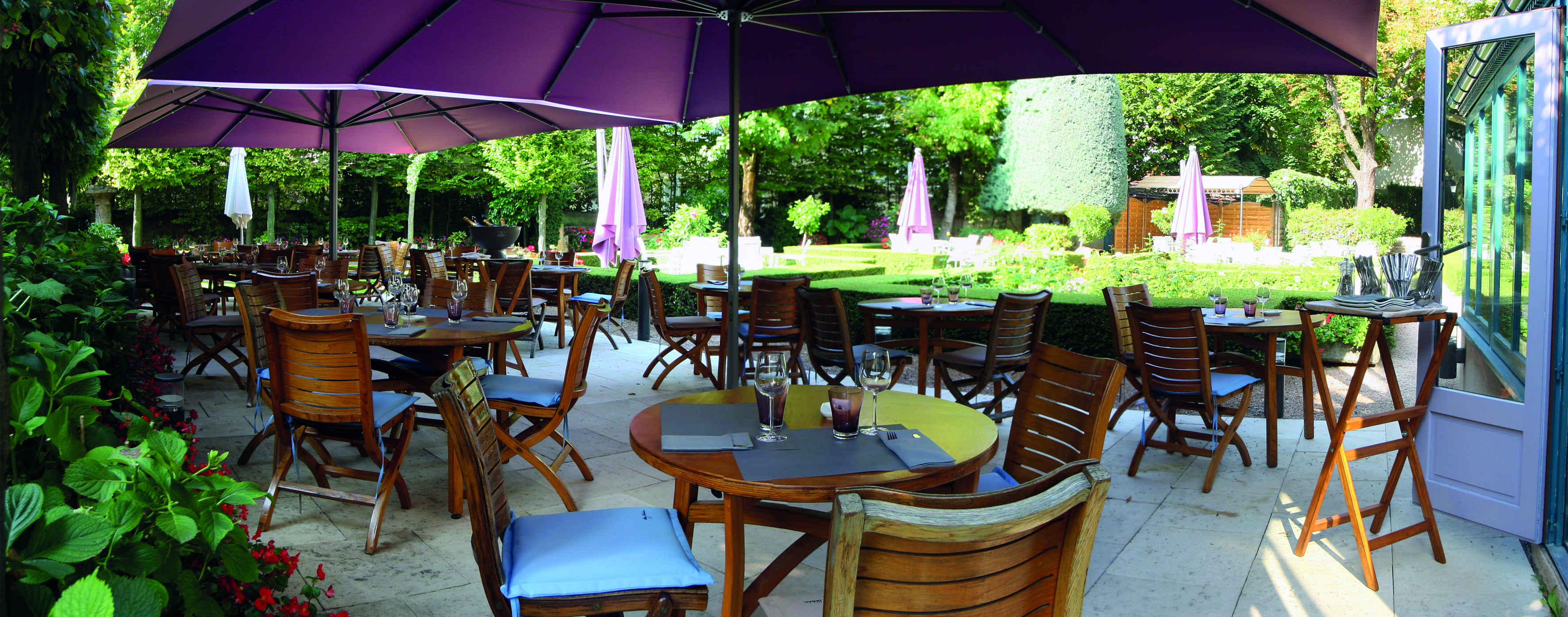 Outdoor terrace seating area with wooden tables and chairs set up around the patio with large purple umbrellas set up 