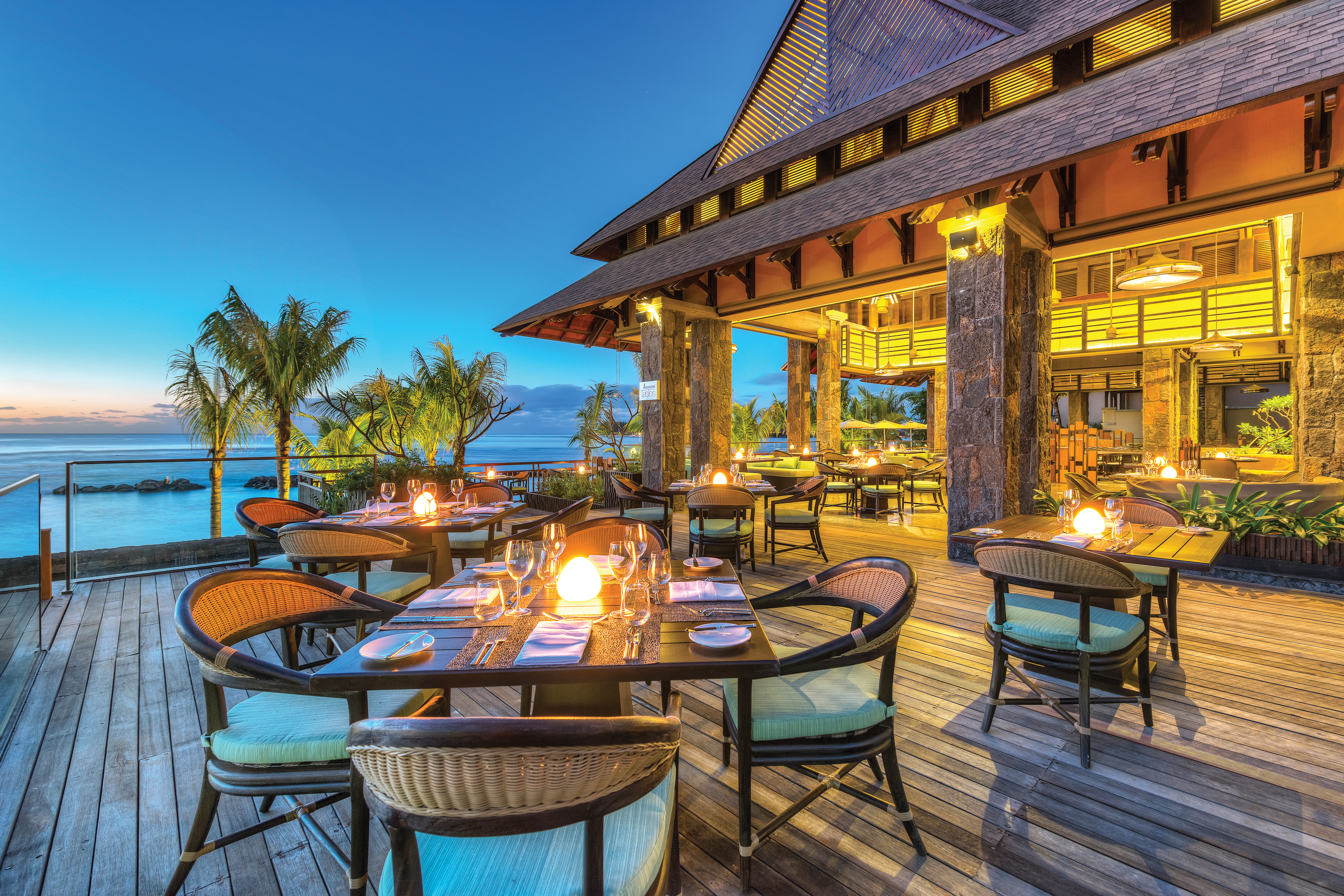 Outdoor dining terrace with candles set up on tables with palm trees and sea in backdrop