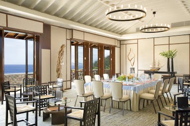 Dining area with tables and chairs set up around the room and doors opening onto an ocean view balcony