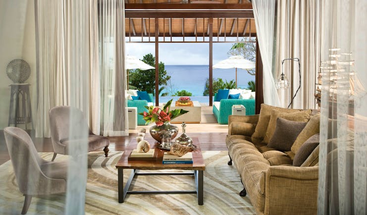 Lounge area with sofa, arm chairs and coffee table with books on and doors opening out onto a pool view