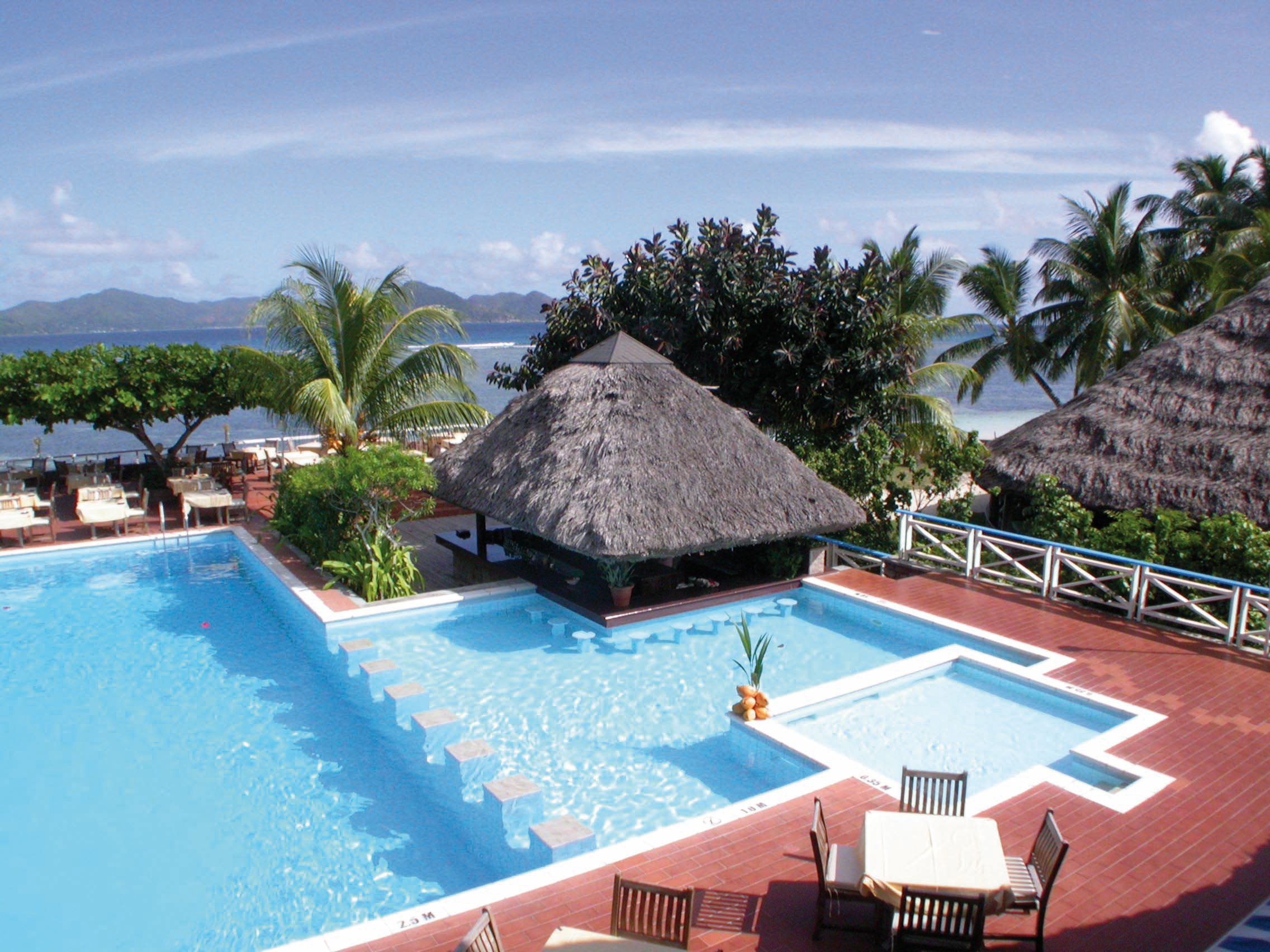 Outdoor swimming pool with palm trees around the edge, looking out over the sea and with beach huts around the pool