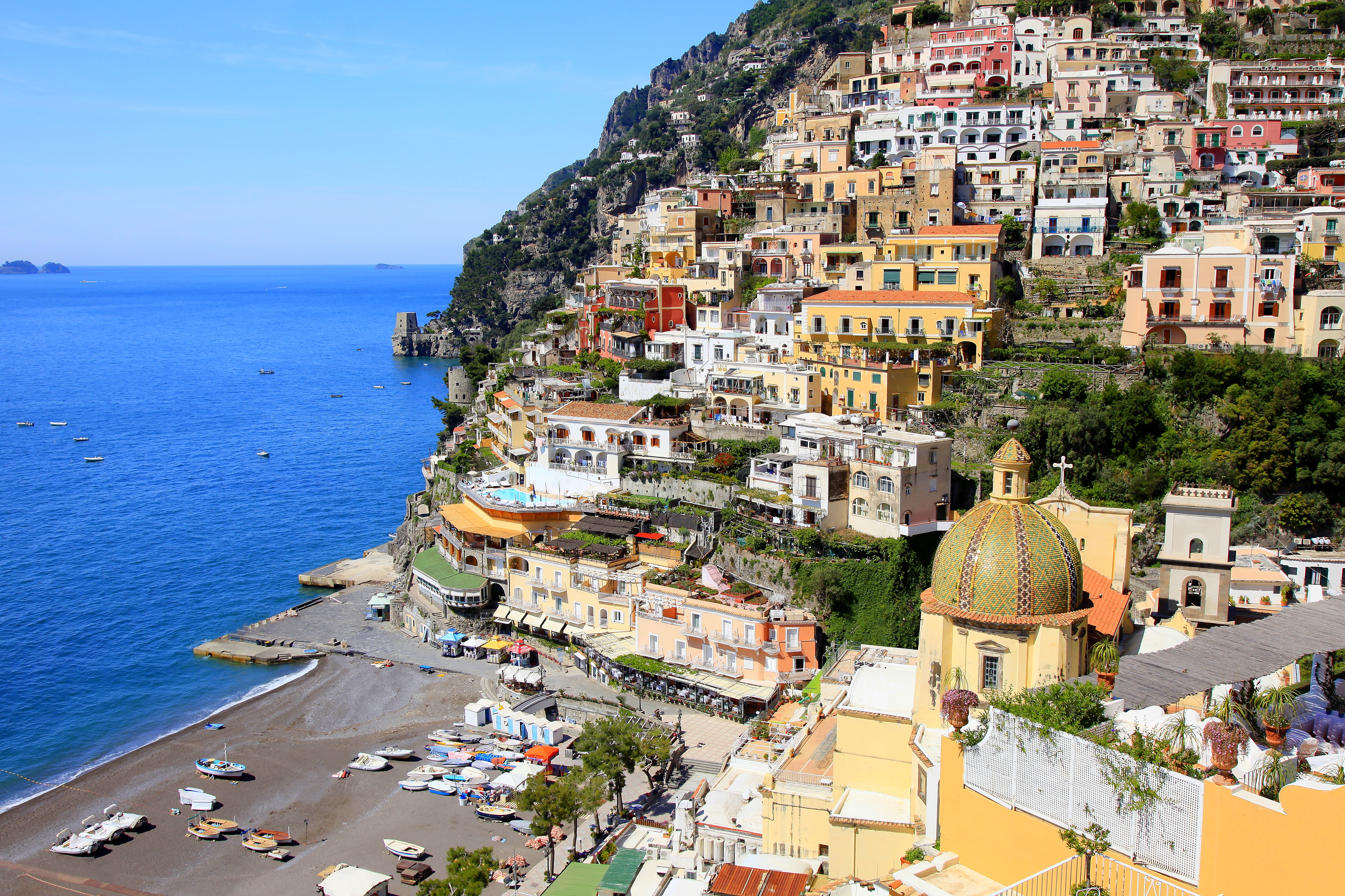 Beach with bright blue sea and dome of church with houses built into the hillside in Positano