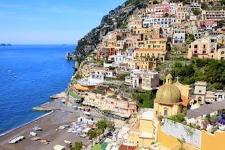 Beach with bright blue sea and dome of church with houses built into the hillside in Positano