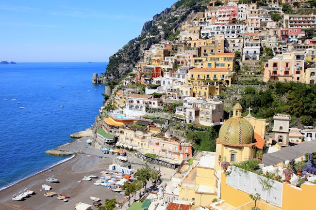 Beach with bright blue sea and dome of church with houses built into the hillside in Positano