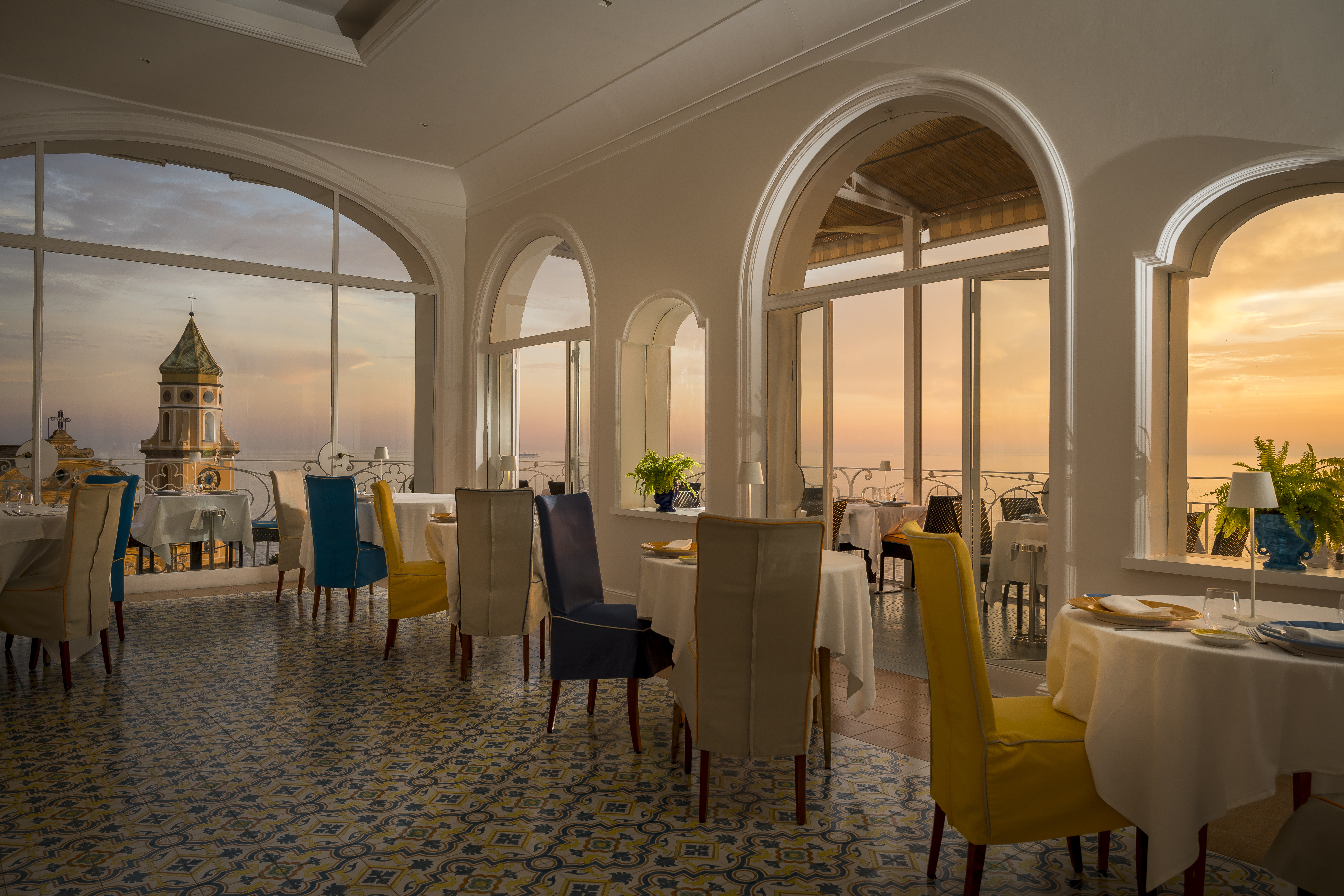 Hotel Tramonto d'Oro Restaurant with elegant blue and yellow chairs set at white-clothed tables, overlooking sunset sea and town views