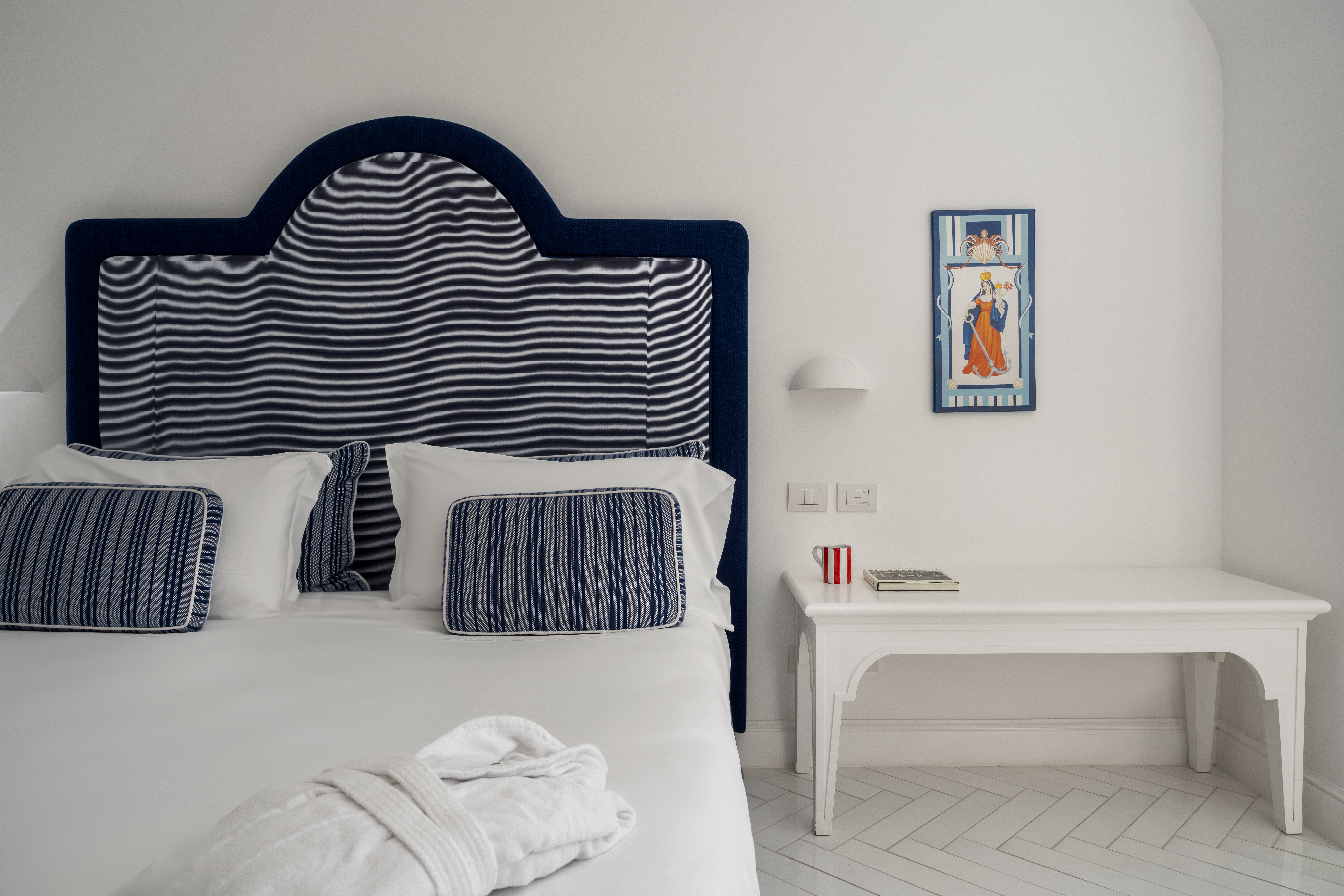 Hotel Tramonto d'Oro guestroom with white walls and sheets, a navy blue headboard and pillows, and traditional Amalfi artwork on the wall