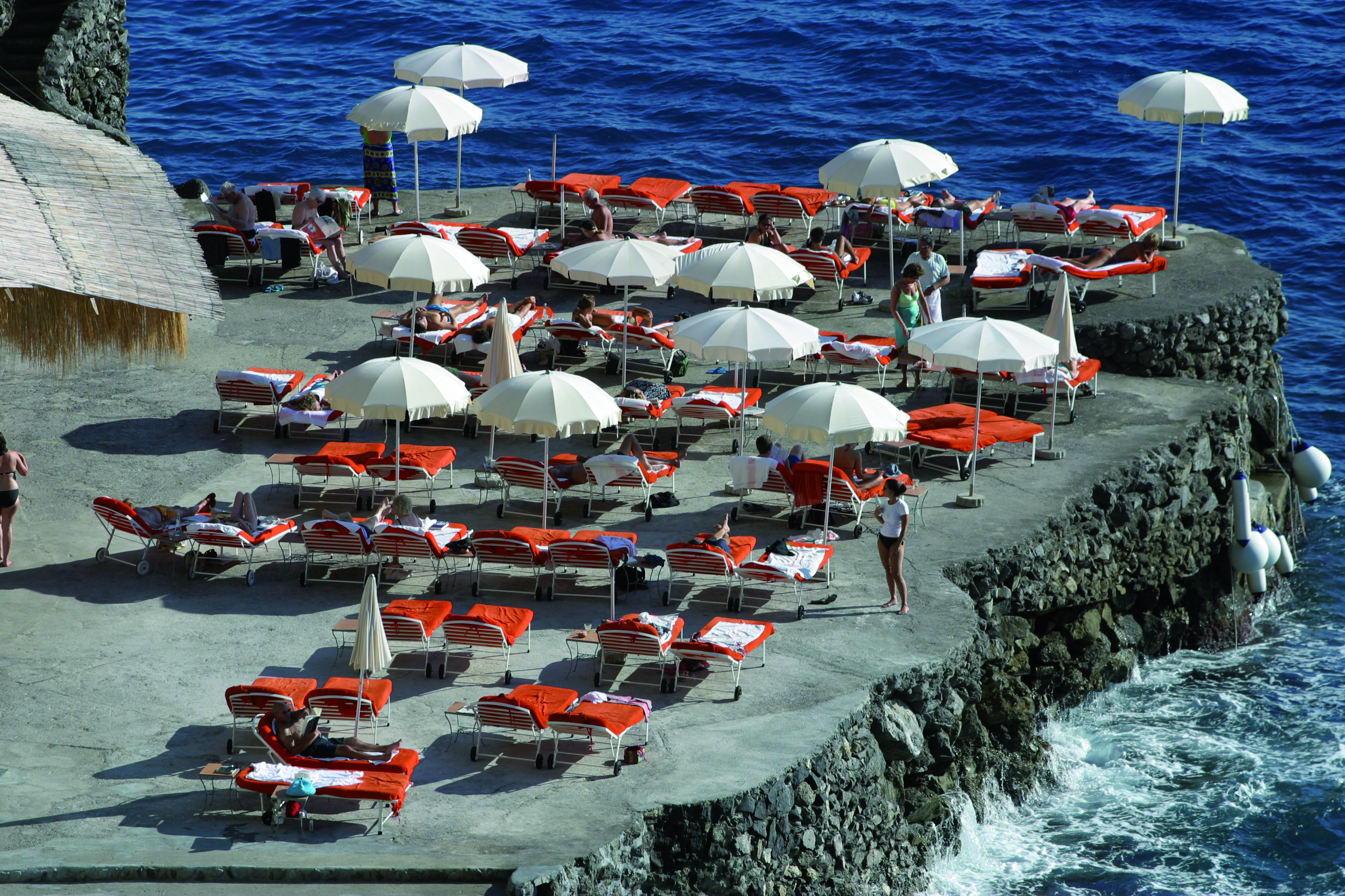 View of the hotel's beach, with red sunbeds and white umbrellas scattered around on the black rocky coast
