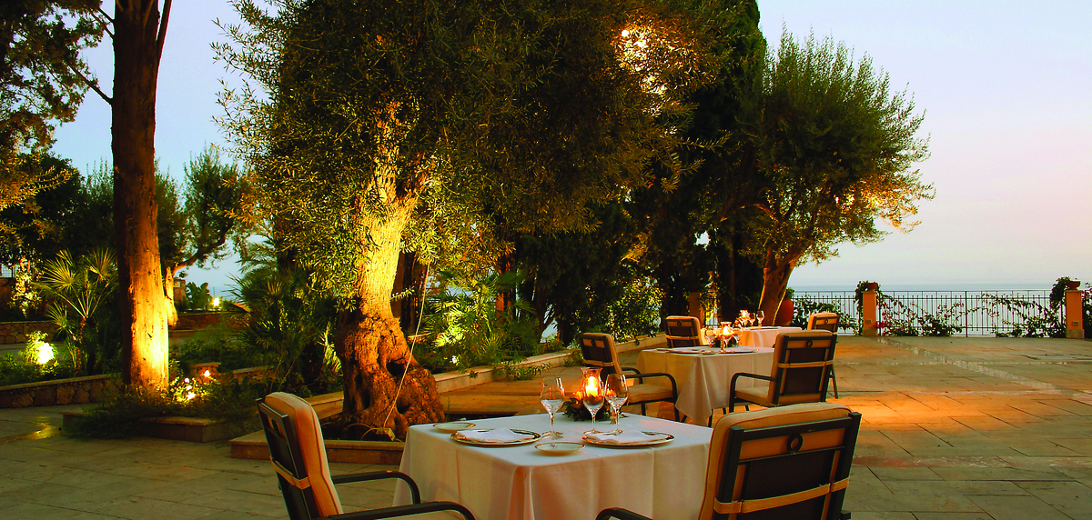 Outdoors restaurant at the Il San Pietro di Positano with palm trees in the background and lights in the trees. A four person table is set 