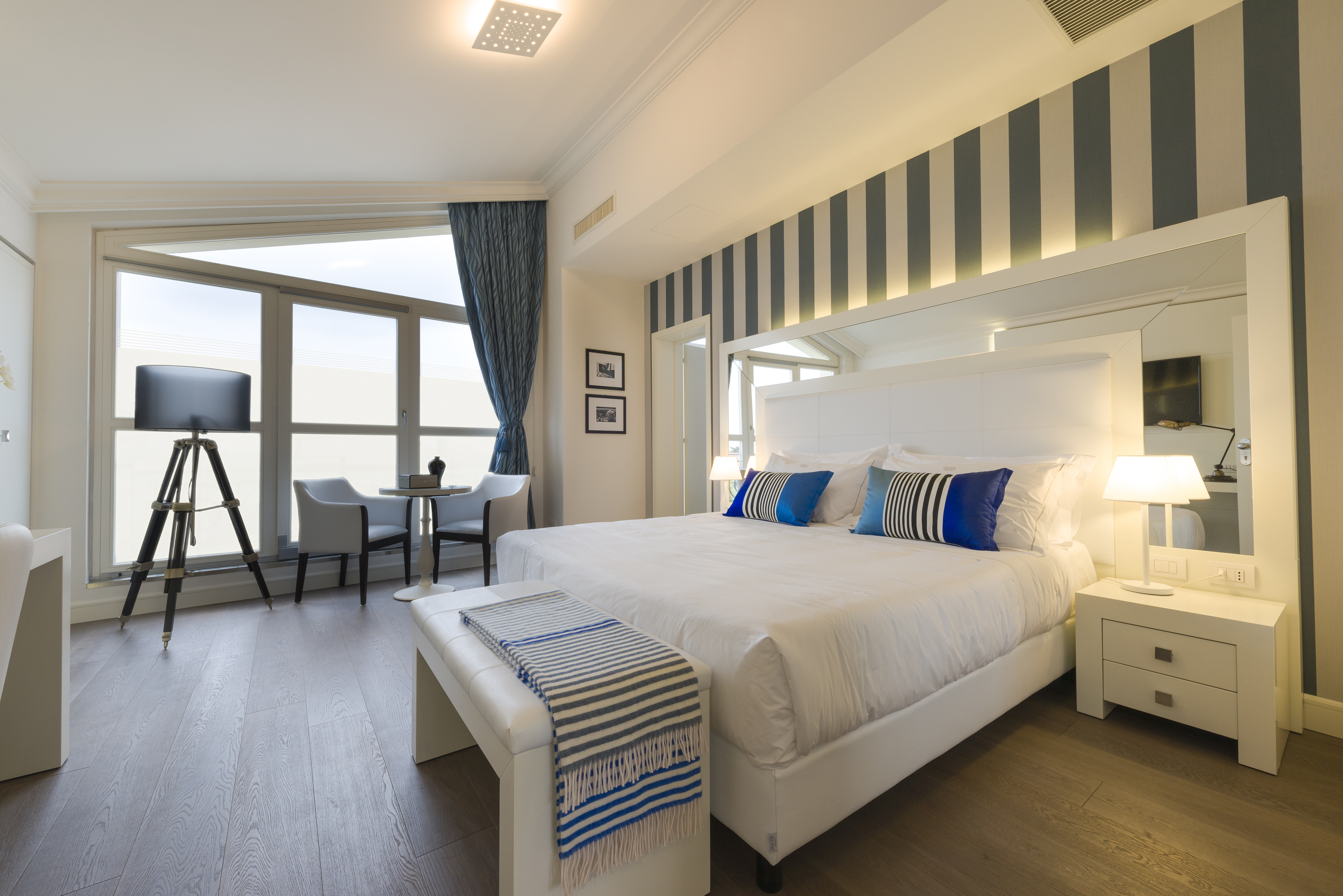 Hotel Palazzo Bezzi room, with white and blue decor, stiped wallpaper around the inset bed, and floor-to-ceiling windows on one wall