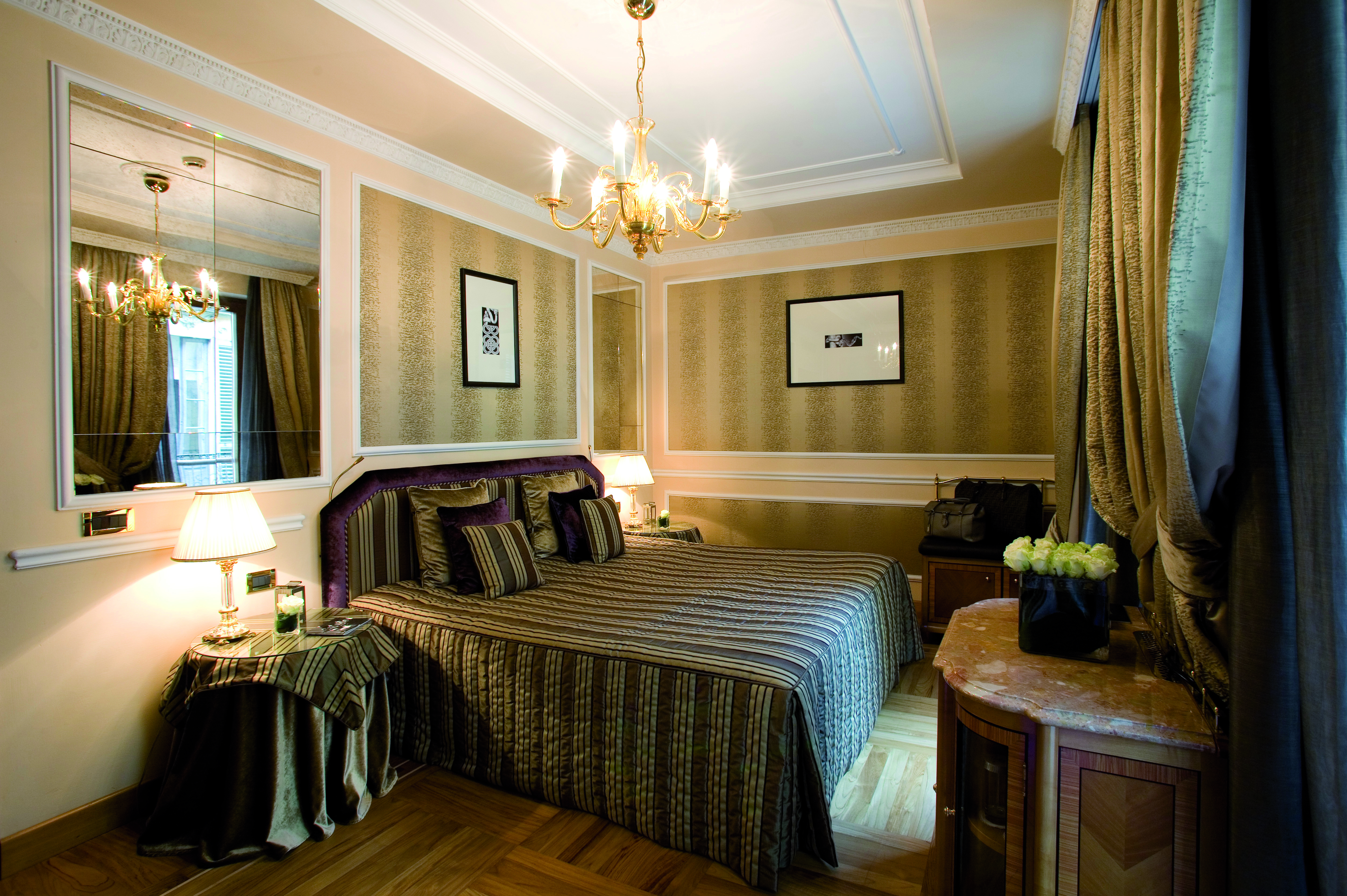 View of a bedroom at the Baglioni Hotel Carlton with a brown and grey colour scheme with a big double bed, mirro and chandelier 