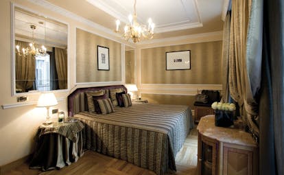 View of a bedroom at the Baglioni Hotel Carlton with a brown and grey colour scheme with a big double bed, mirro and chandelier 