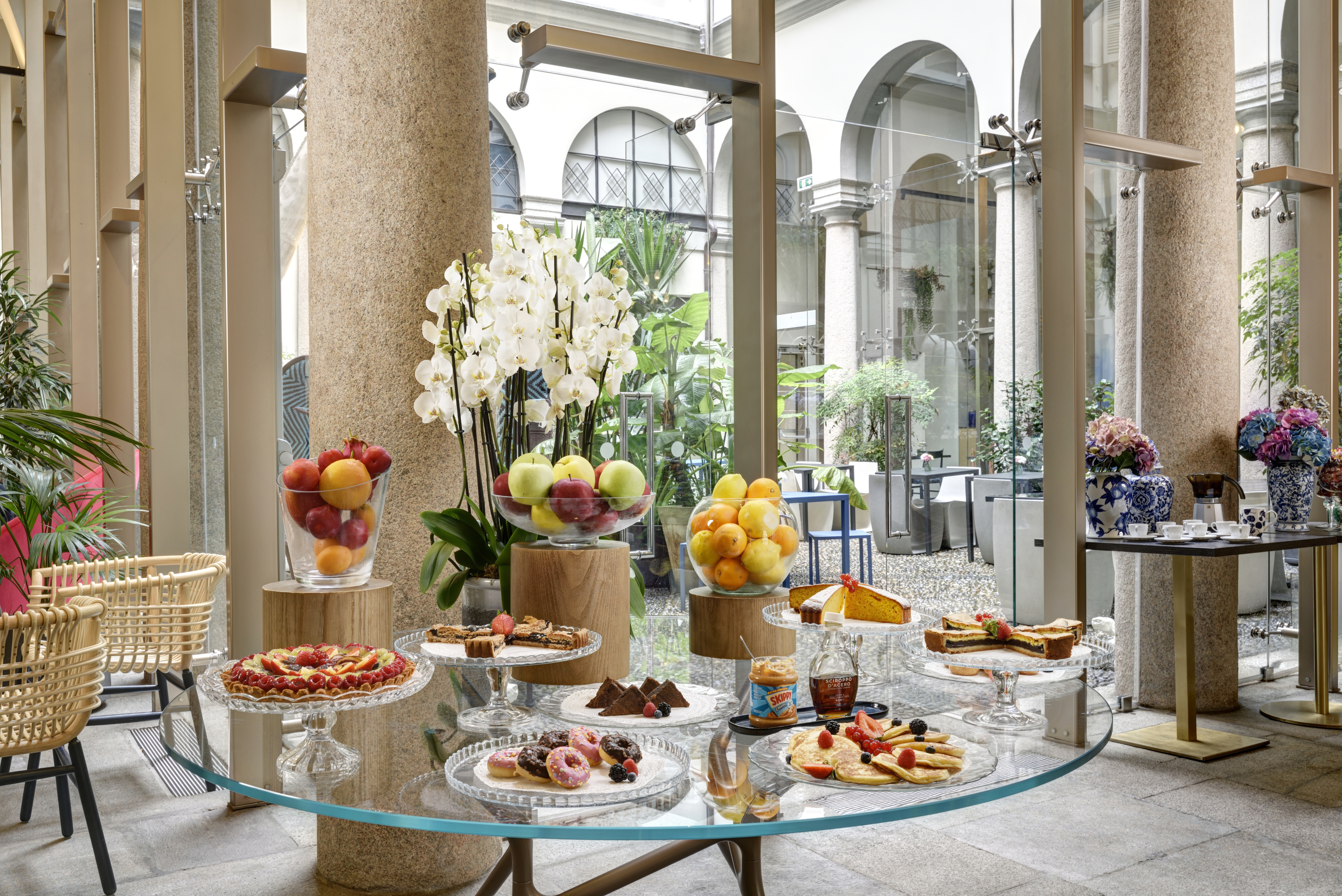 Hotel Indigo Milan breakfast buffet, served on round glass tables with flower and fruit centrepieces, with the bright courtyard and arched windows visible in the background