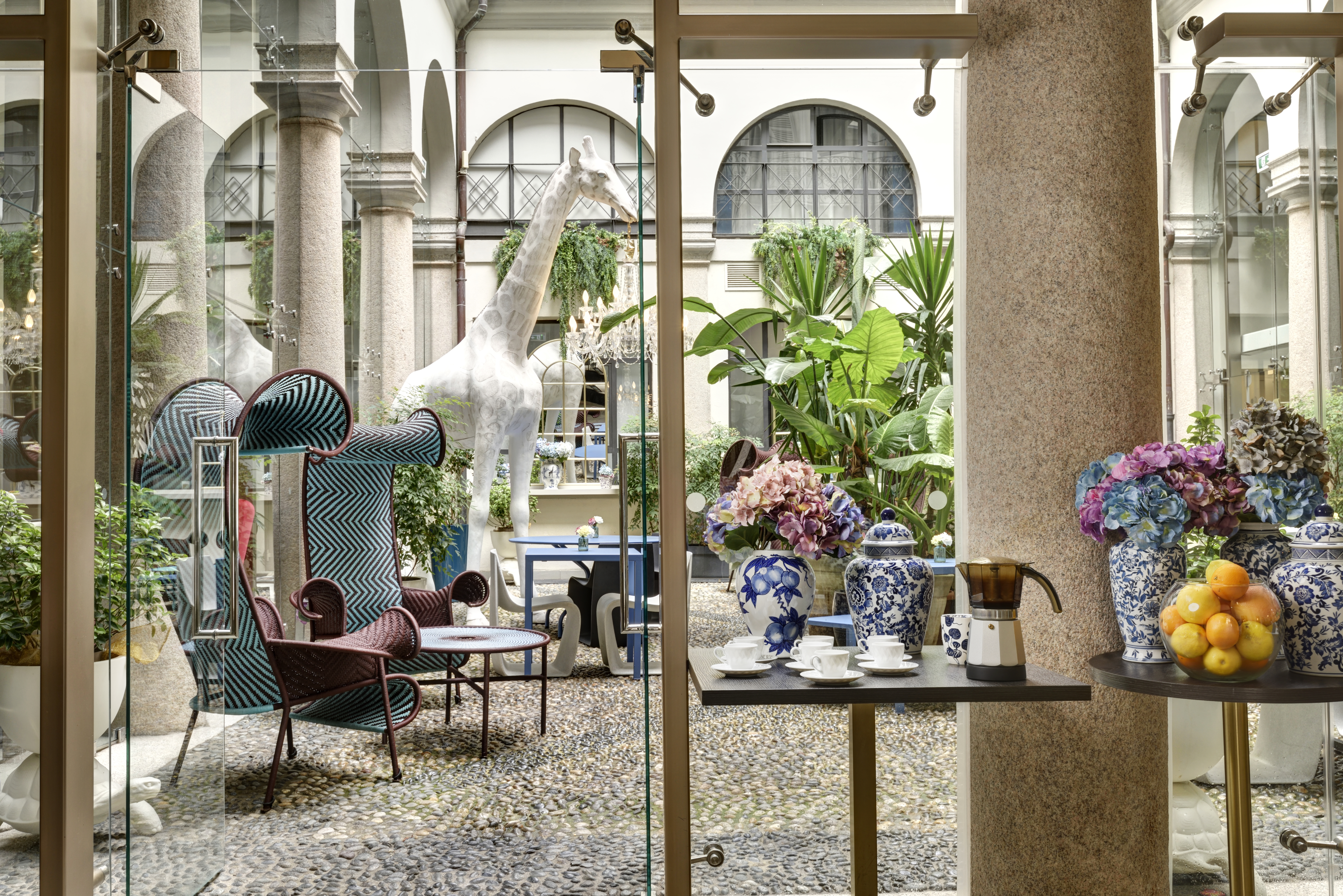 Hotel Indigo Milan courtyard, with tall arched windows surrounding it, high-backed modern chairs, tall leafy palms, and a lifesize white giraffe statue