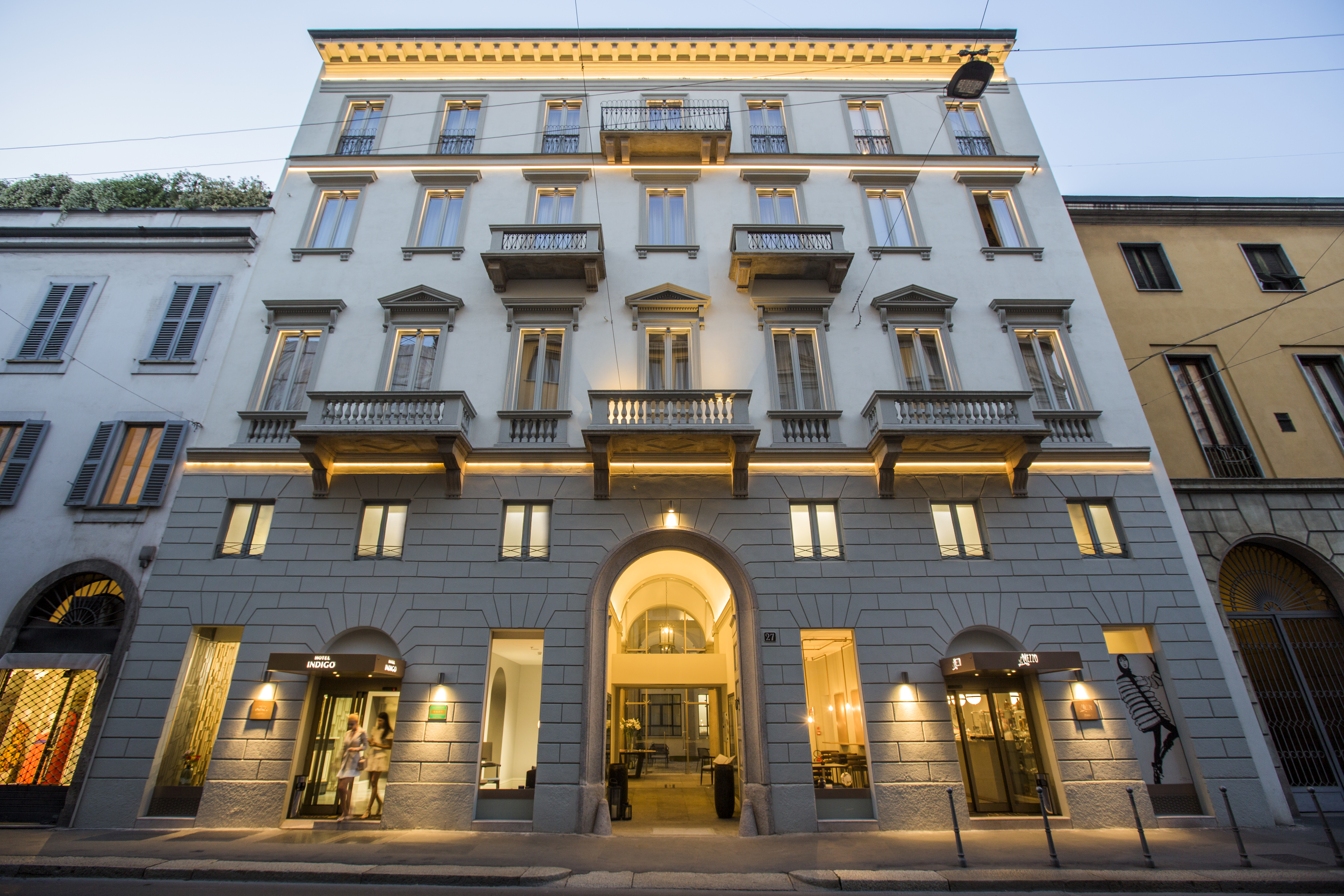 Hotel Indigo Milan exterior, showing white and stone walls, ornate window frames, and down-lit arches on the ground floor