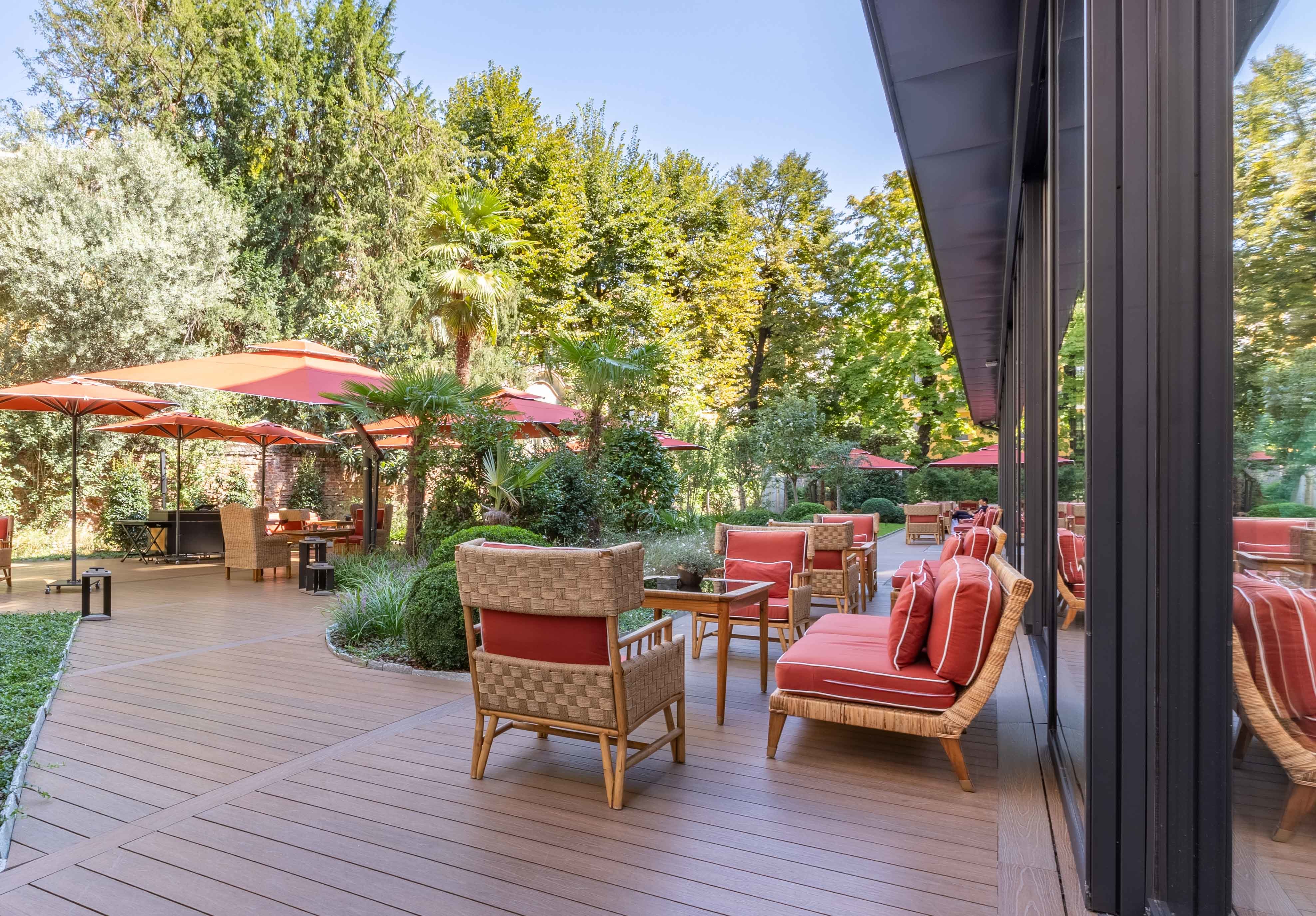 Hotel Portrait Milano garden terrace with red sofas and parasols set amidst tall trees and collections of bushes