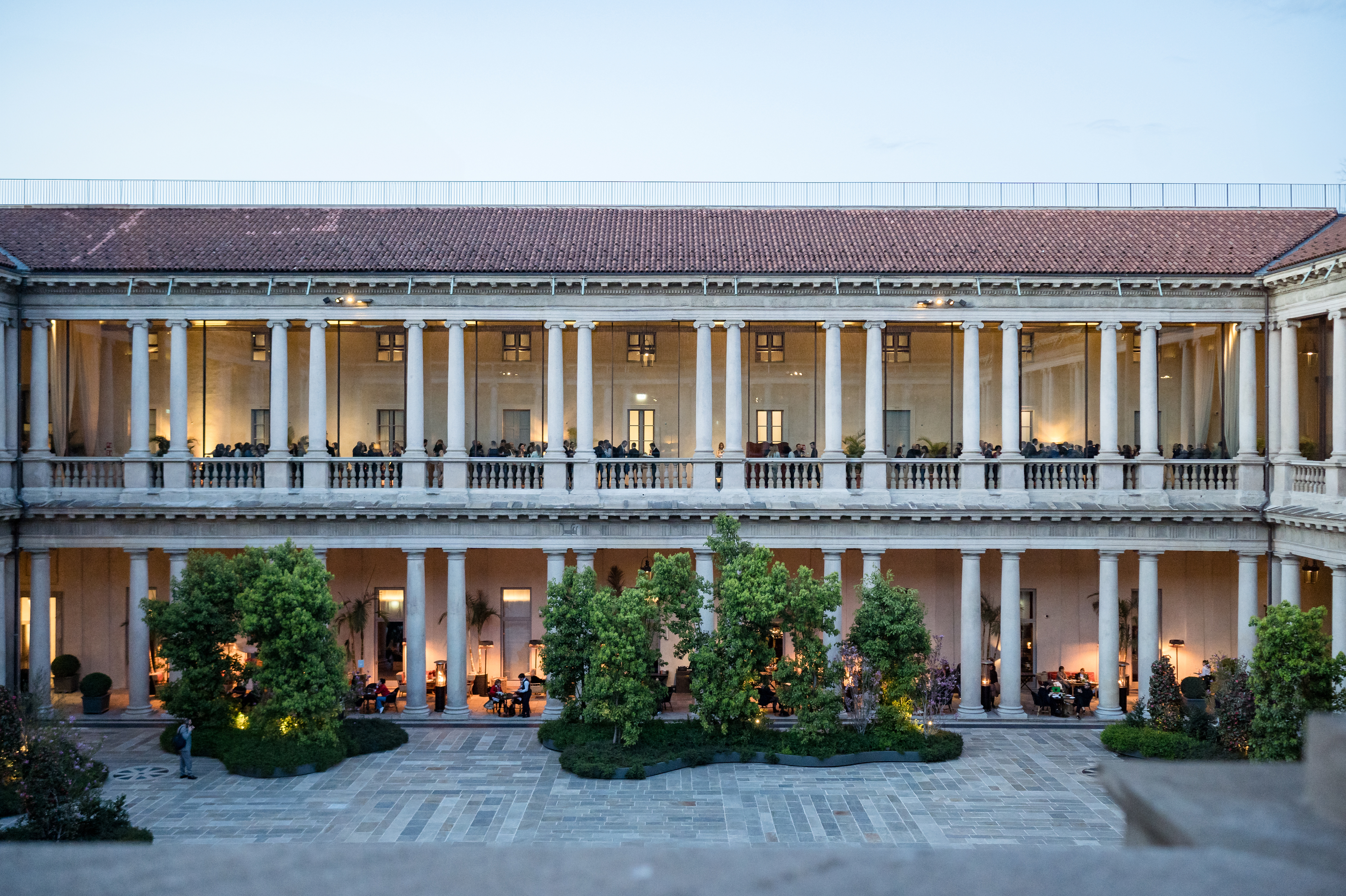 Hotel Portrait Milano internal courtyard, showing two levels of hotel building with interior balconies lined with pillars. Some trees line the edges of the piazza