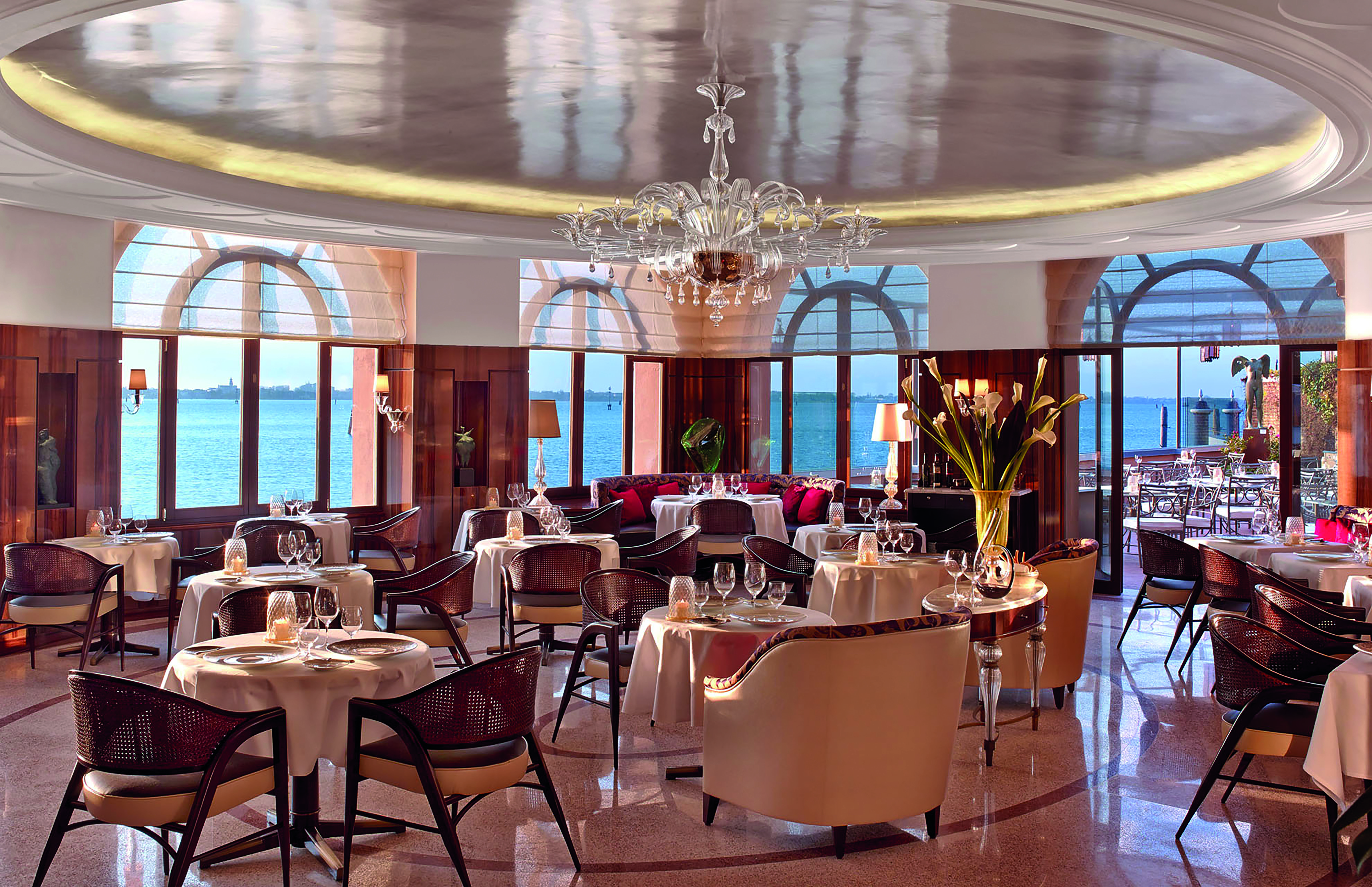 View of the restaurant in the Belmond Hotel Cipriani with a red and orange colour scheme with big arching windows round the circumference of the room with sea views