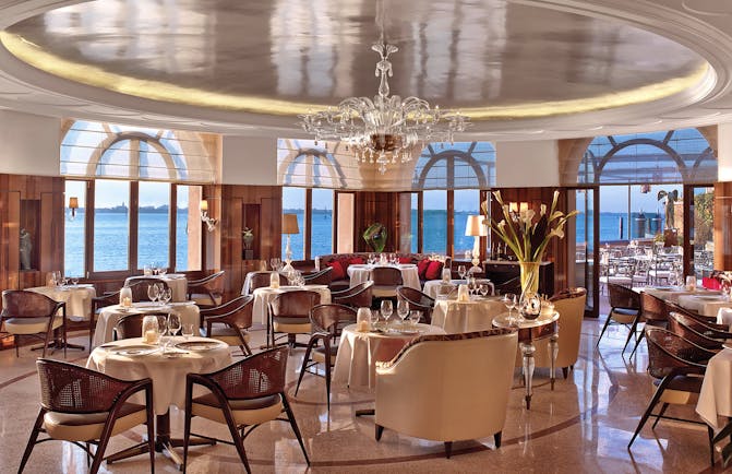 View of the restaurant in the Belmond Hotel Cipriani with a red and orange colour scheme with big arching windows round the circumference of the room with sea views