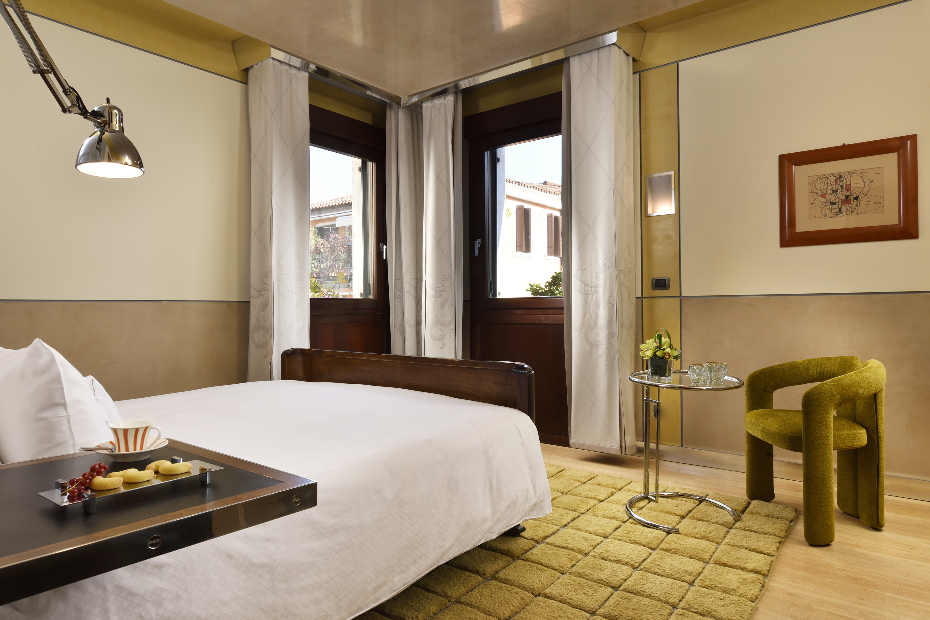 Ca' Pisani Deluxe Room with plush gold rug on wooden floor, double bed with white linen, and two windows in the corner of the room looking out over Venice views