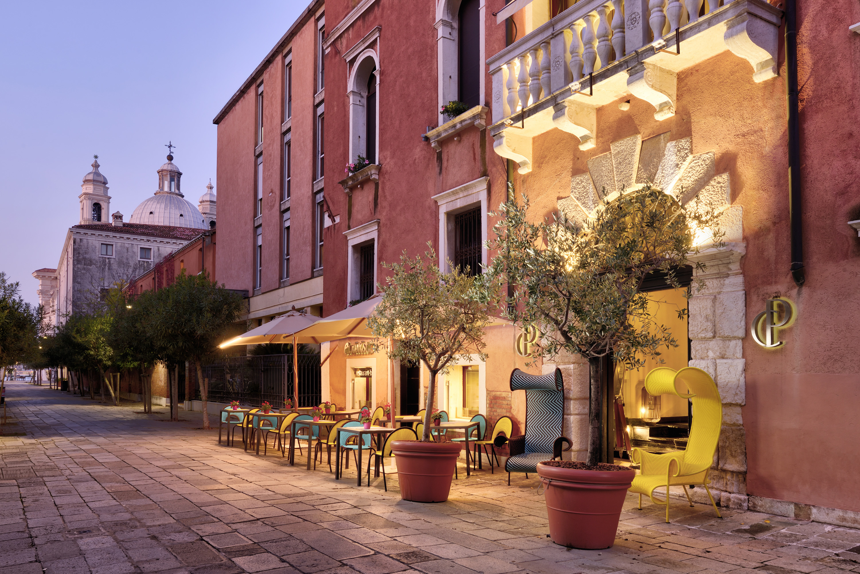 Ca' Pisani exterior view, showing pink-washed hotel walls, with stone-framed arches, potted trees, outdoor seating, and the rooftops of Venice at evening time