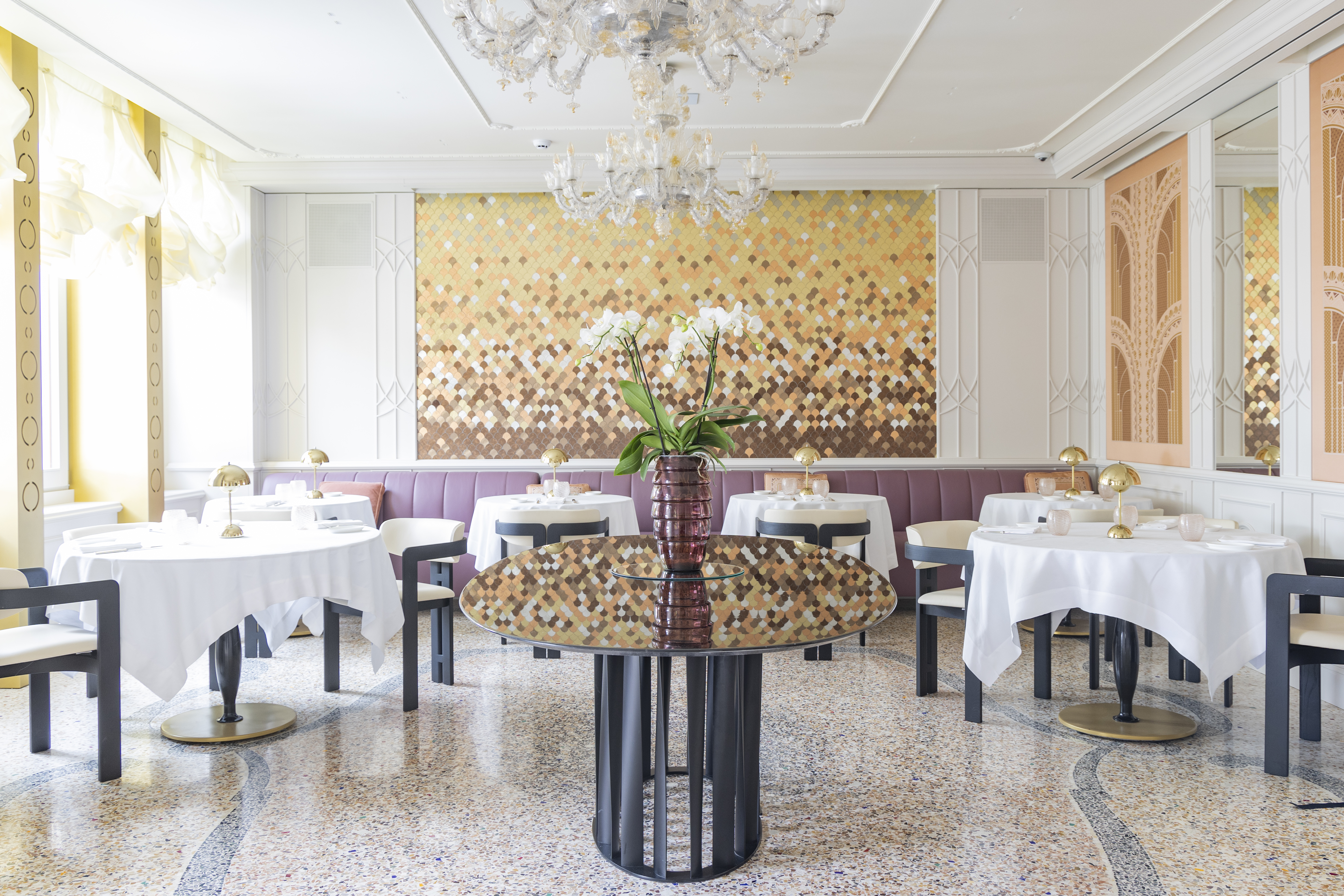 Londra Palace Venezia LPV Restaurant with round white-clothed tables arranged around a floral centrepiece, with mosaic-style artworks on the walls and large bright windows on the left
