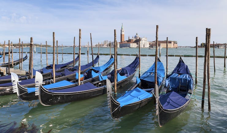 Row of gondolas with blue covers moored to posts opposite San Giorgio Maggiore Venice