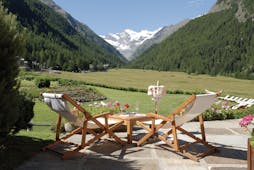 Terrace area with wooden deckchairs laid out, overlooking a green lawn and the italian alps, with snow-topped mountains far away 