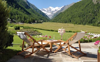 Terrace area with wooden deckchairs laid out, overlooking a green lawn and the italian alps, with snow-topped mountains far away 