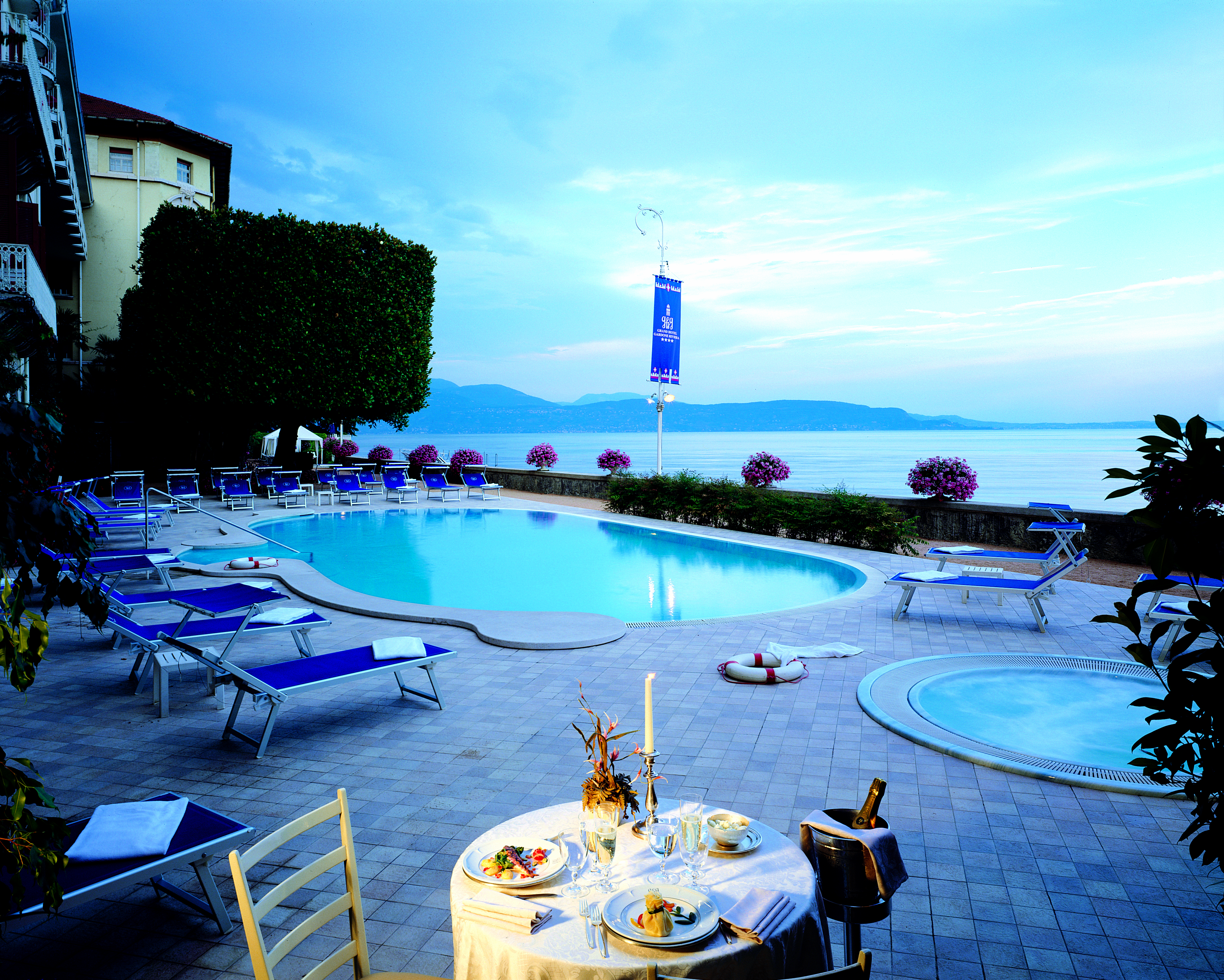 Grand Hotel Gardone view of the pool with blue sun beds set up surrounding it and a circular plunge poor on the side with the lake in the background